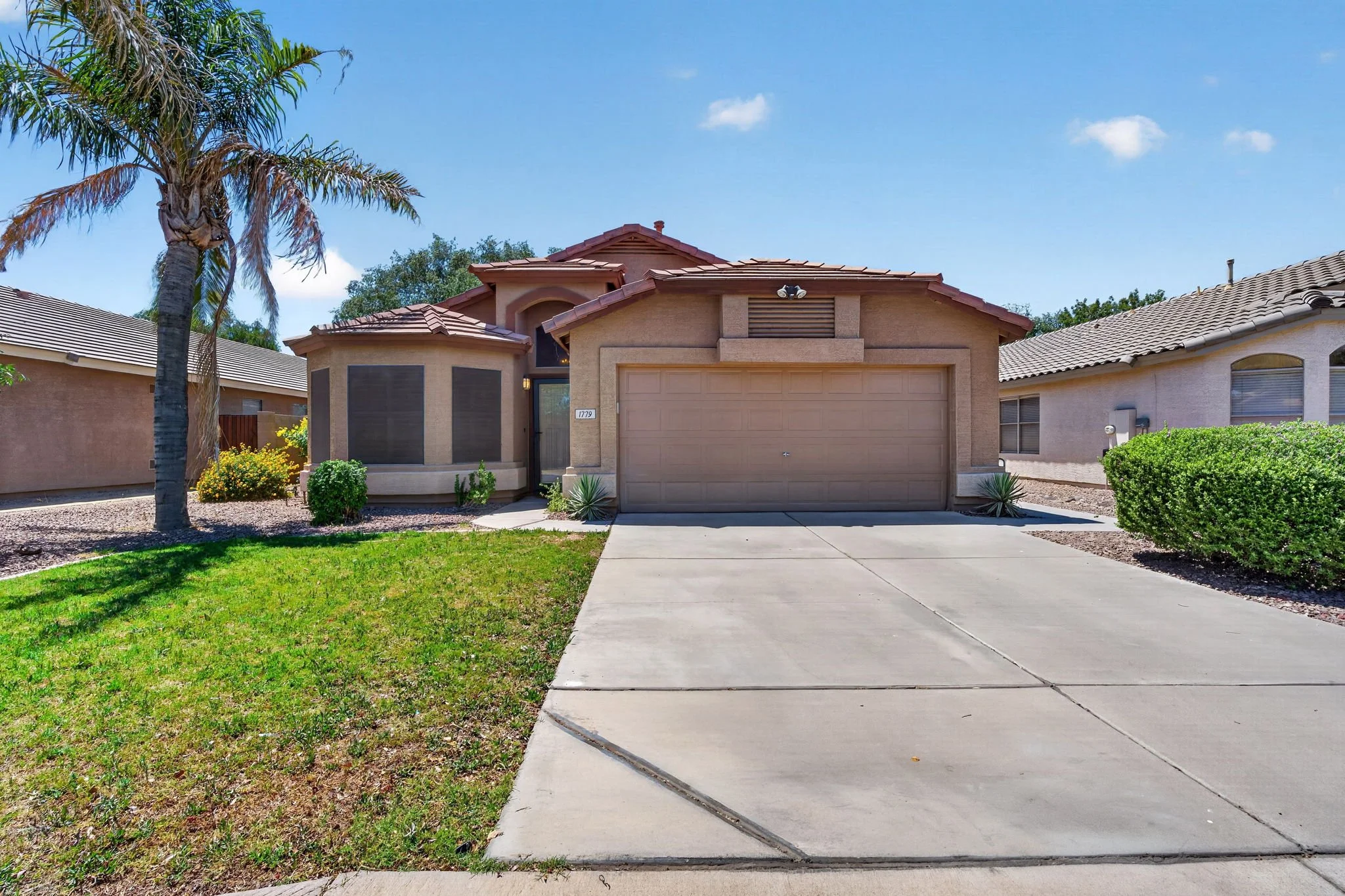 A suburban house with a front yard, palm tree, concrete driveway, and a beige garage door under a partly cloudy blue sky.