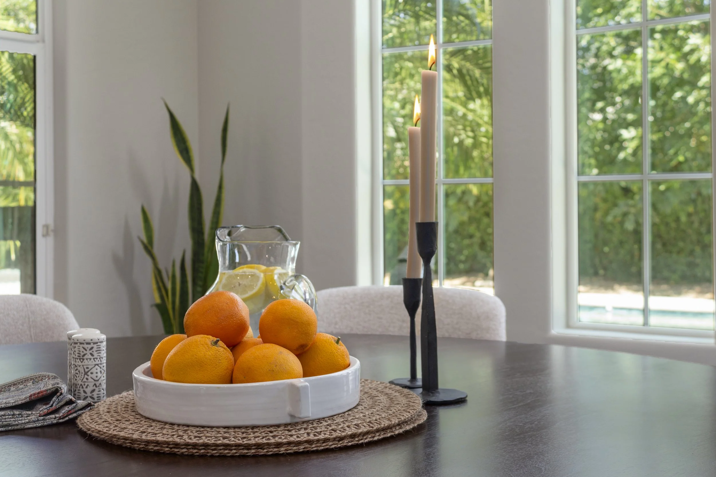 A dining table with a white tray of oranges, a glass pitcher of lemon water with lemon slices, a decorative candle holder, and a candle holder with three lit candles. In the background, there are large windows with a view of green trees and a tall pl