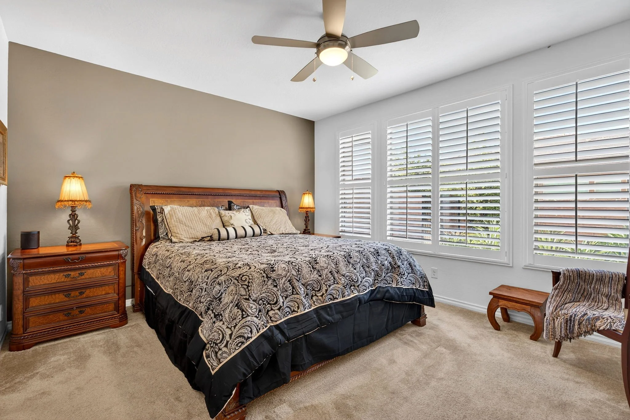 A bedroom with a wooden bed frame, patterned bedspread, and matching pillows. There are two bedside tables with lamps, a wooden chair with patterned upholstery, a ceiling fan, large windows with white shutters, and beige carpet flooring.