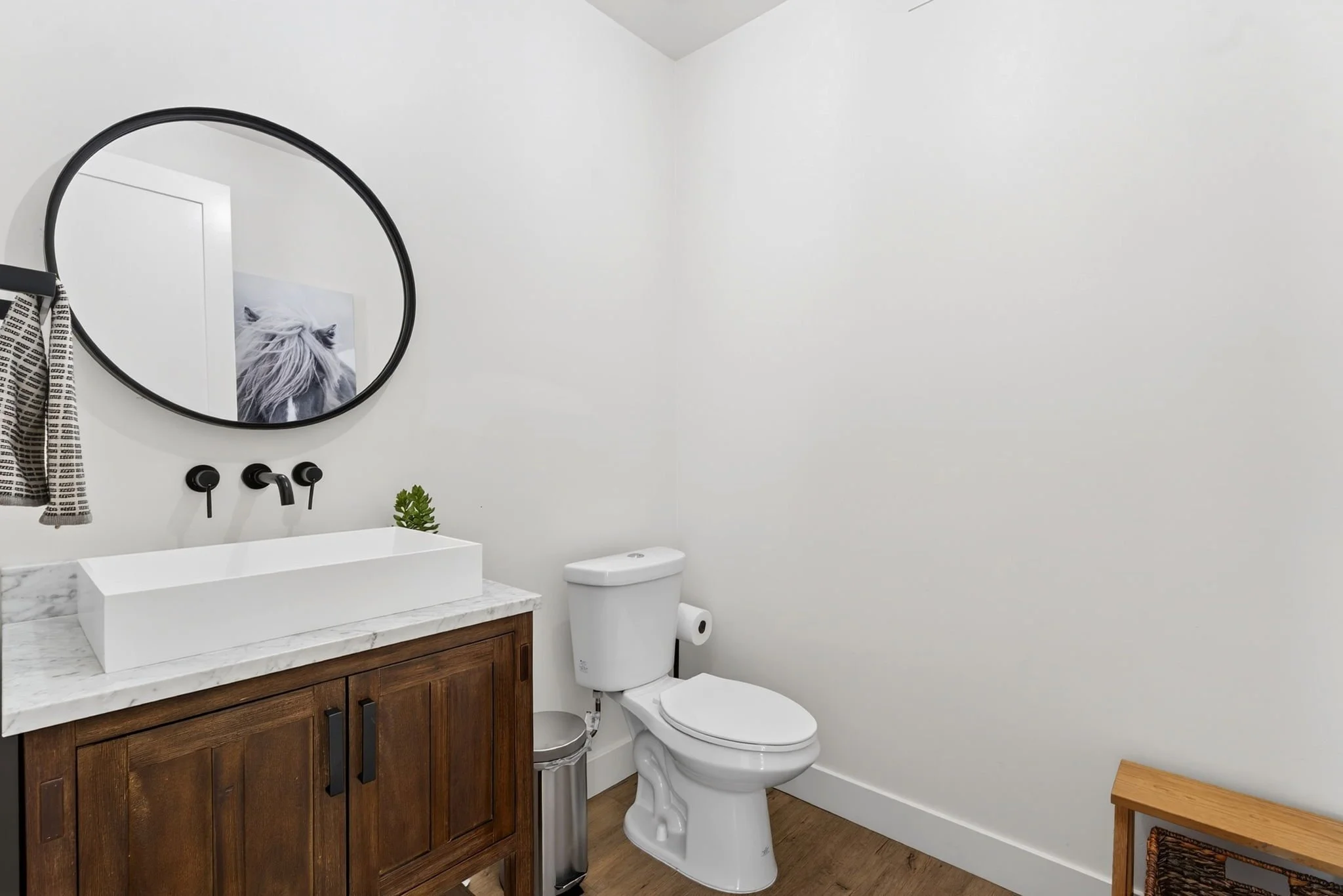 A modern bathroom with a white toilet, a wooden vanity with a white marble countertop, a rectangular vessel sink, a round black-framed mirror, black faucet fixtures, a small green plant on the countertop, a toilet paper roll on the holder, and a smal