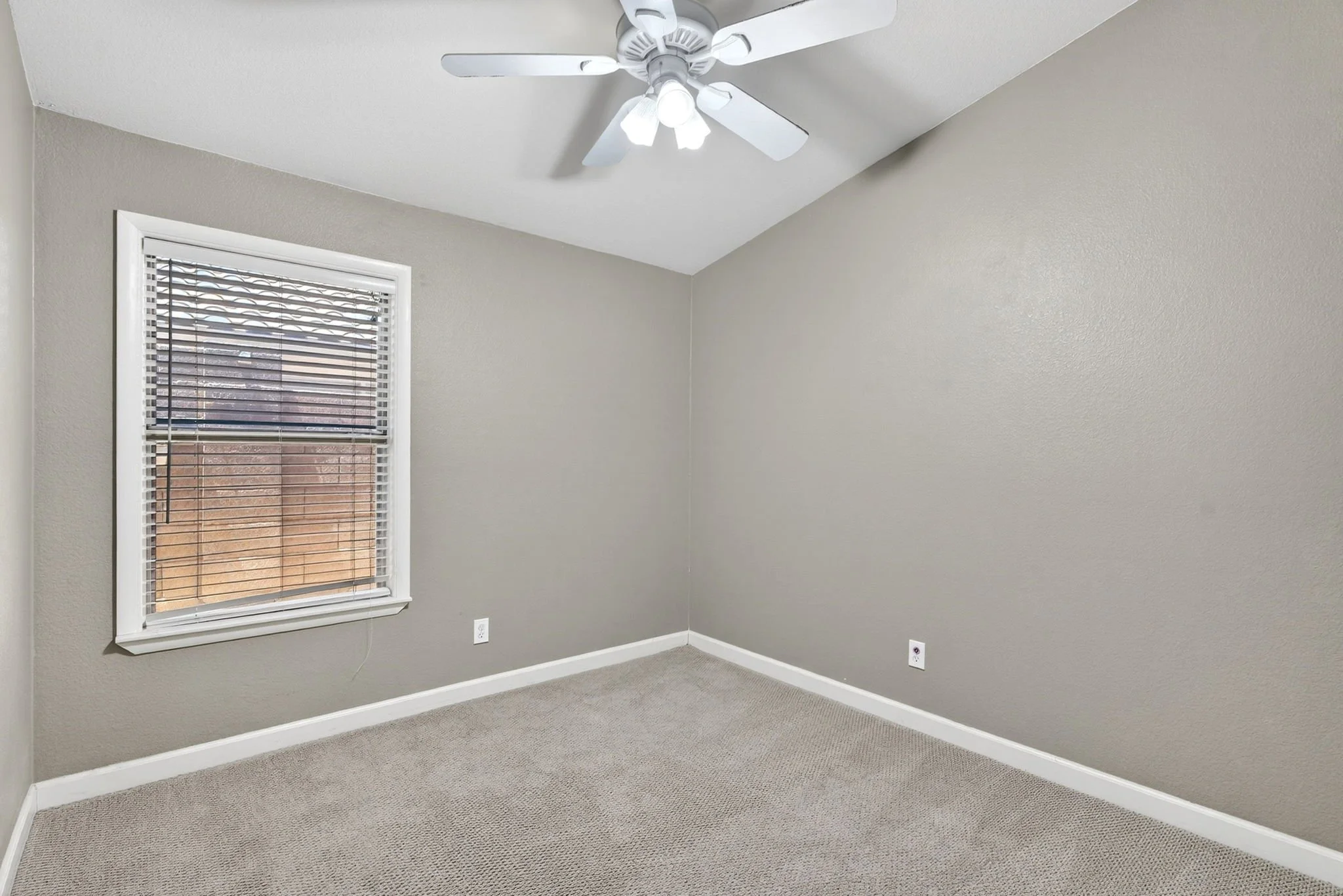 Empty room with beige walls, a window with blinds, a ceiling fan with lights, electric outlets, and carpeted flooring.
