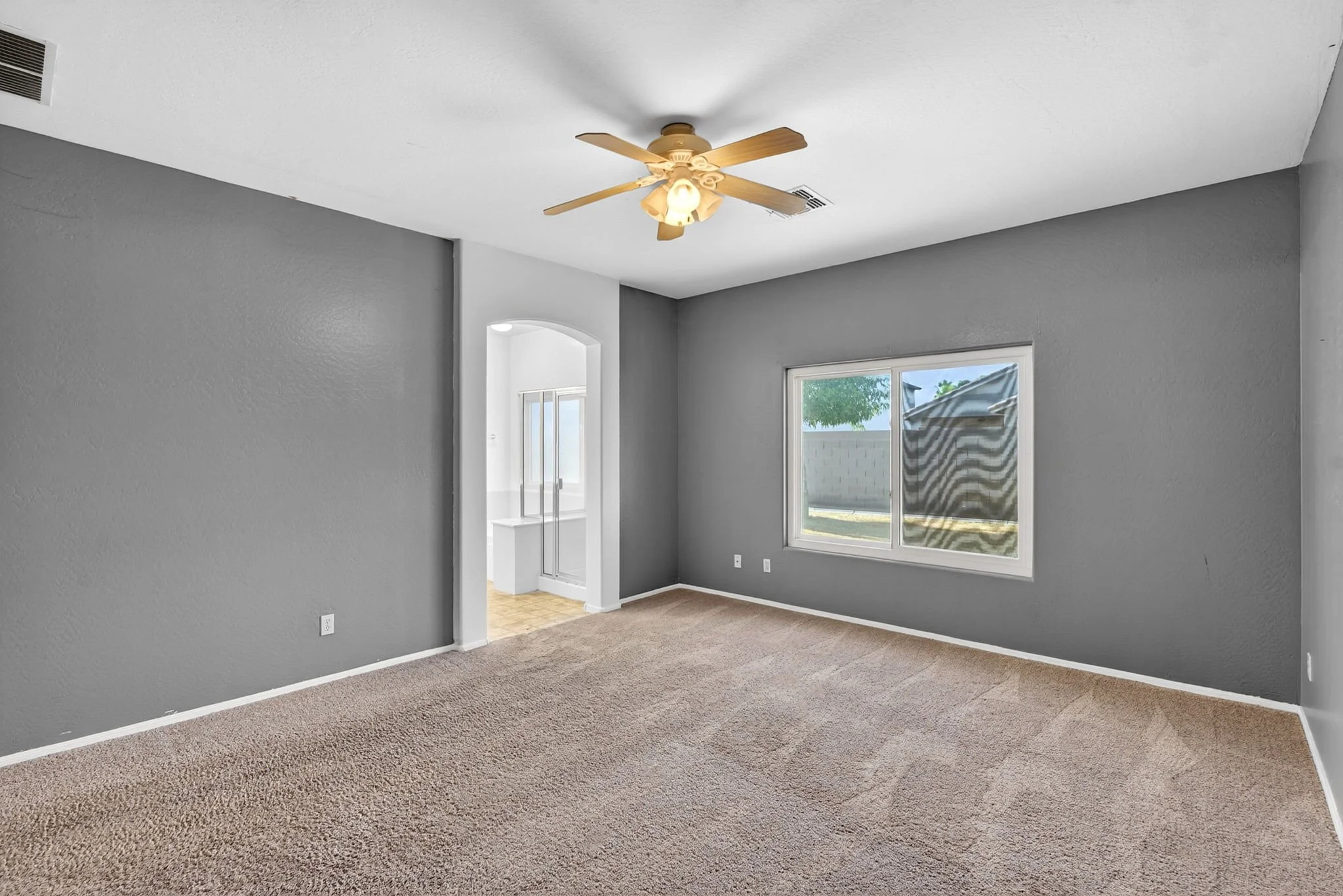Empty living room with light brown carpet, gray walls, ceiling fan, and sliding glass door leading outside.