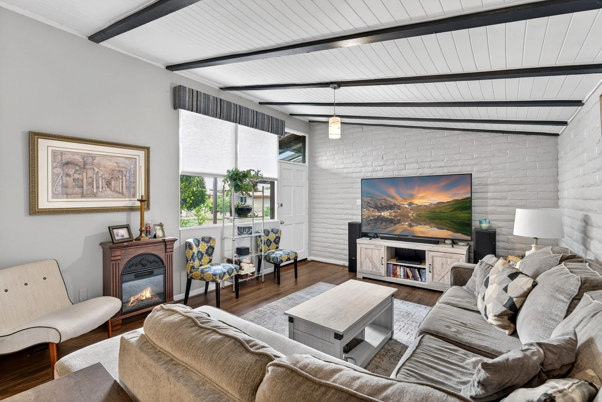 Living room with beige sofas, a white brick accent wall, a large flat-screen TV, framed artwork, chairs near a window, and wood flooring.