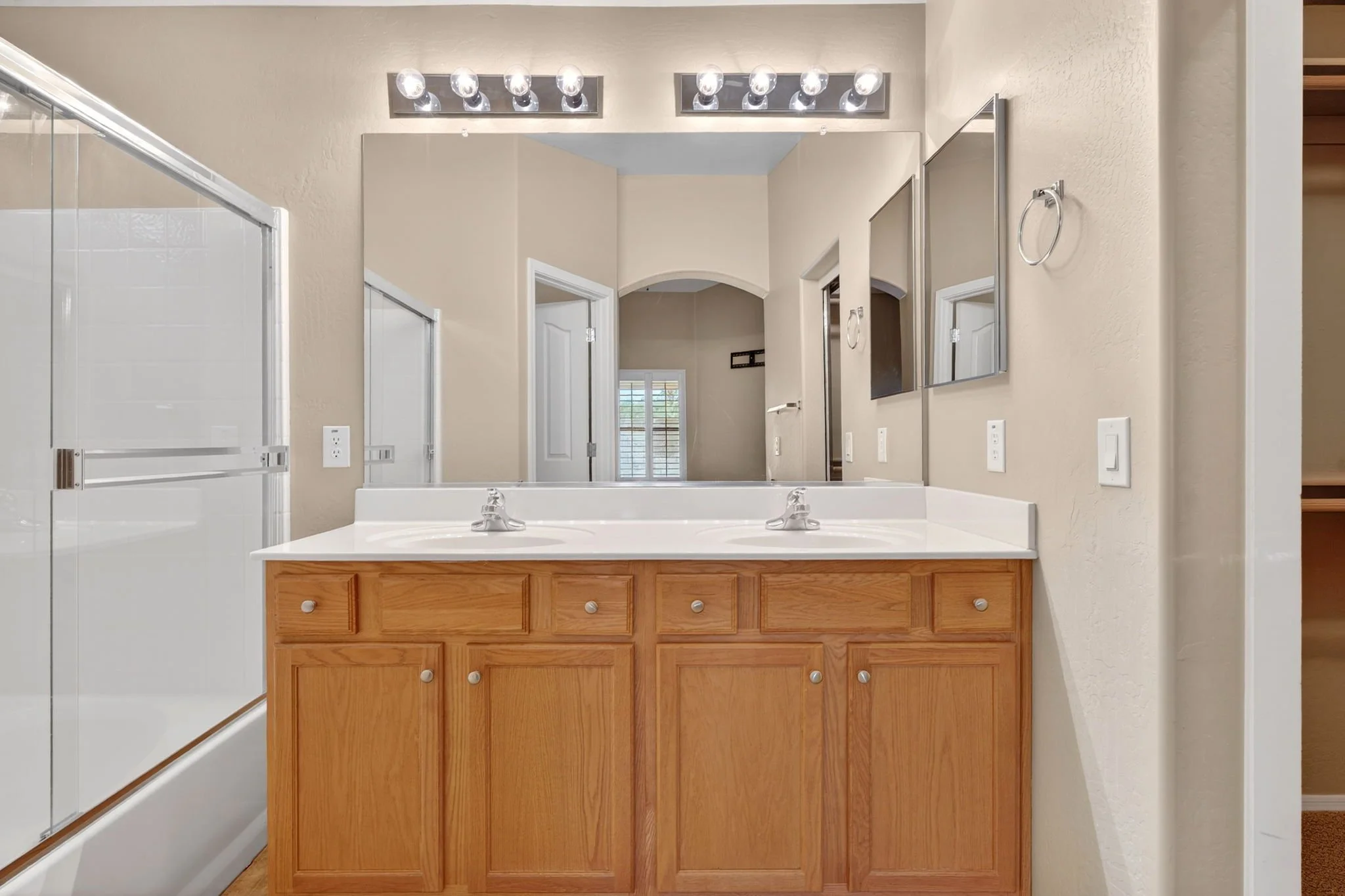 Bathroom with a large mirror, two sinks, a wooden cabinet, a glass shower door on the left, beige walls, and light fixtures above the mirror.