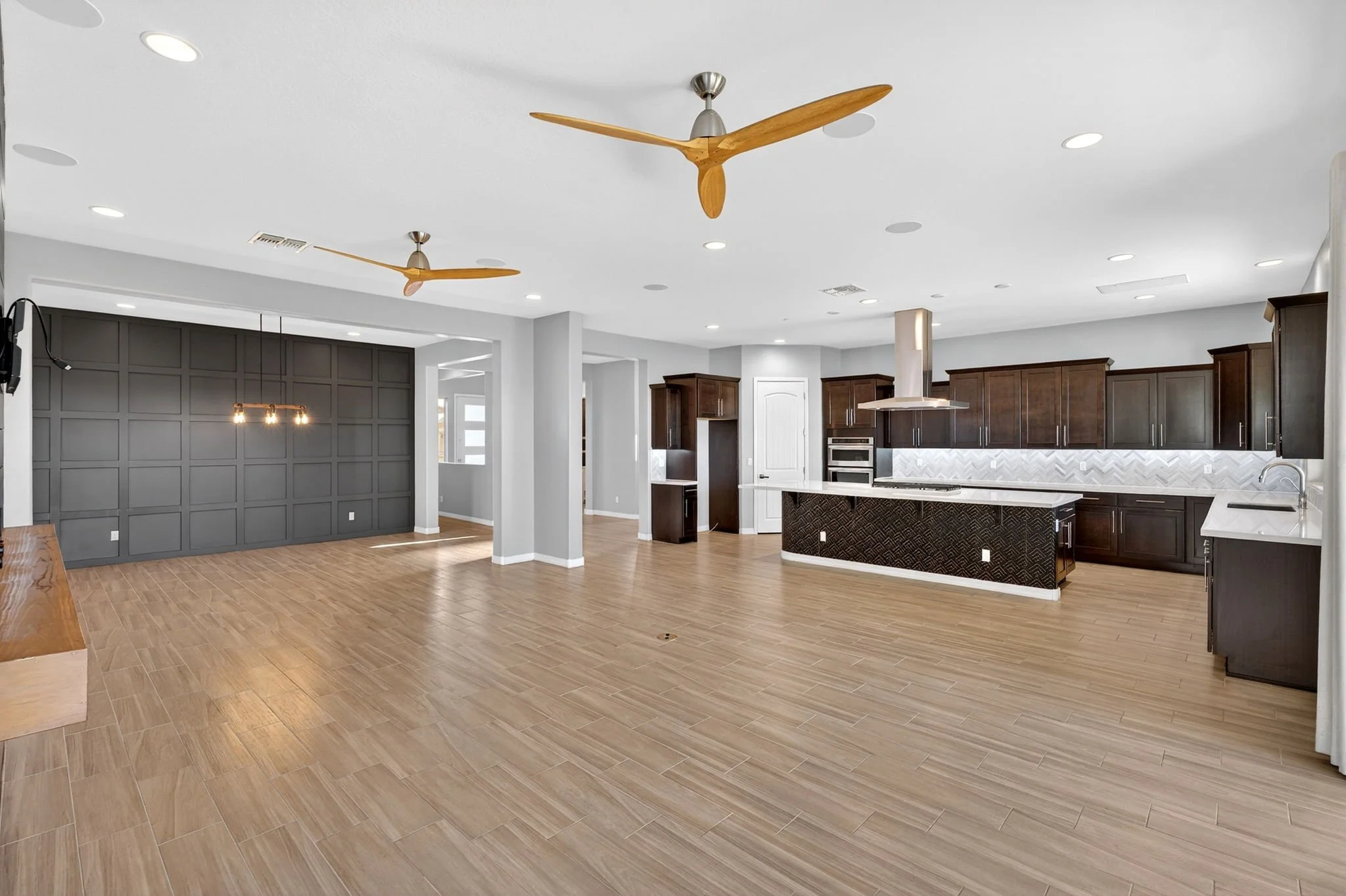 Modern open-concept kitchen and living area with wood floors, dark wood cabinets, a white island, and ceiling fans with wooden blades.