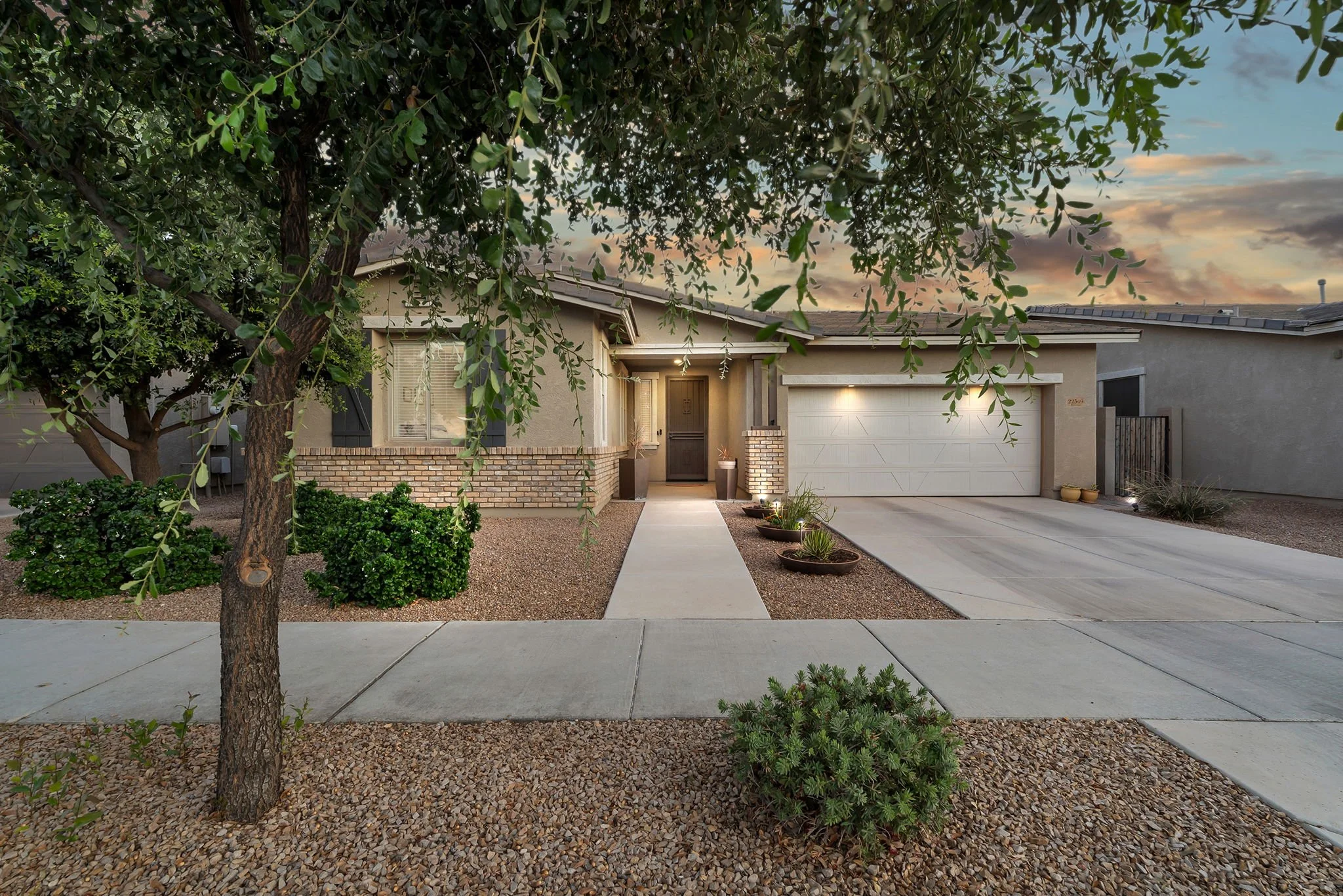 A single-family house with a brick and stucco exterior, a front yard with gravel and a concrete pathway, a large tree partially obscuring the house, and a cloudy sunset sky in the background.