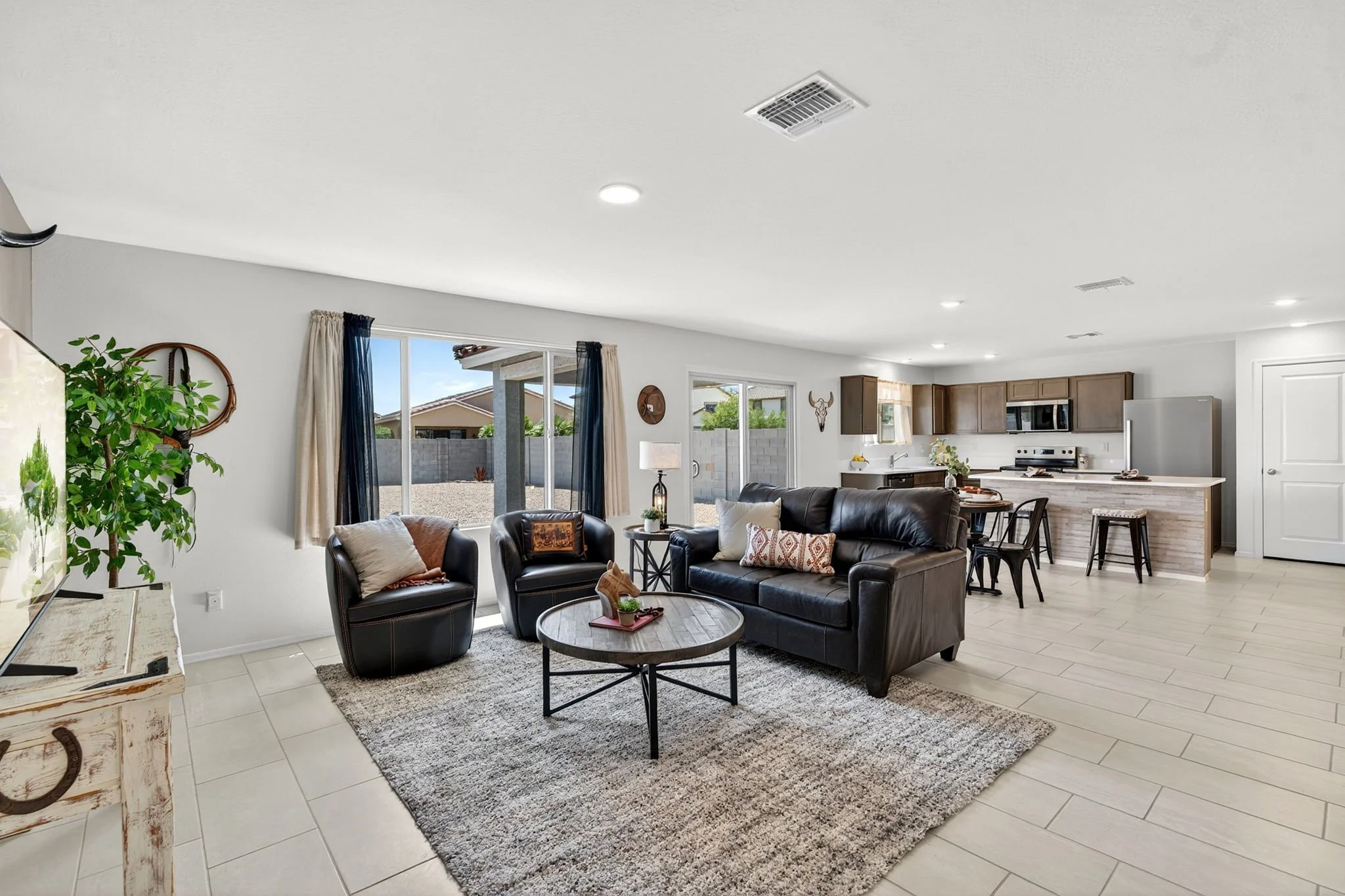 Bright open-concept living room with beige tiled flooring, black leather sofa, matching armchair, round coffee table, area rug, large window with curtains, and a view of backyard. Visible kitchen with wooden cabinets, stainless steel appliances, and 