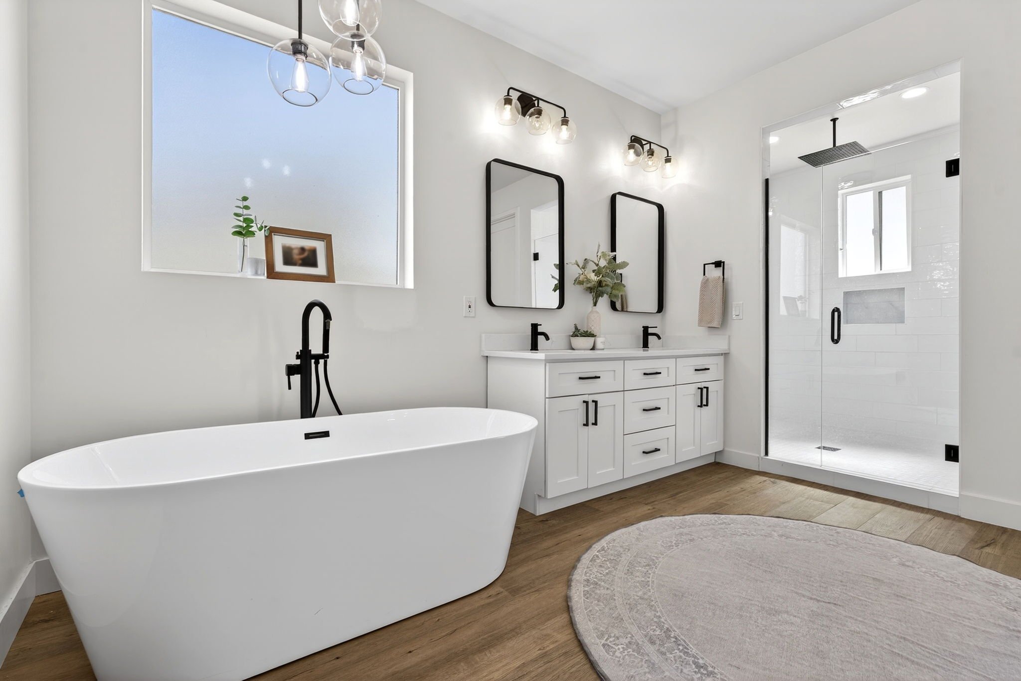 Modern bathroom with a white freestanding bathtub, a double vanity with black fixtures, two black-framed mirrors, a shower area with glass door, and decorative lighting fixtures on white walls.