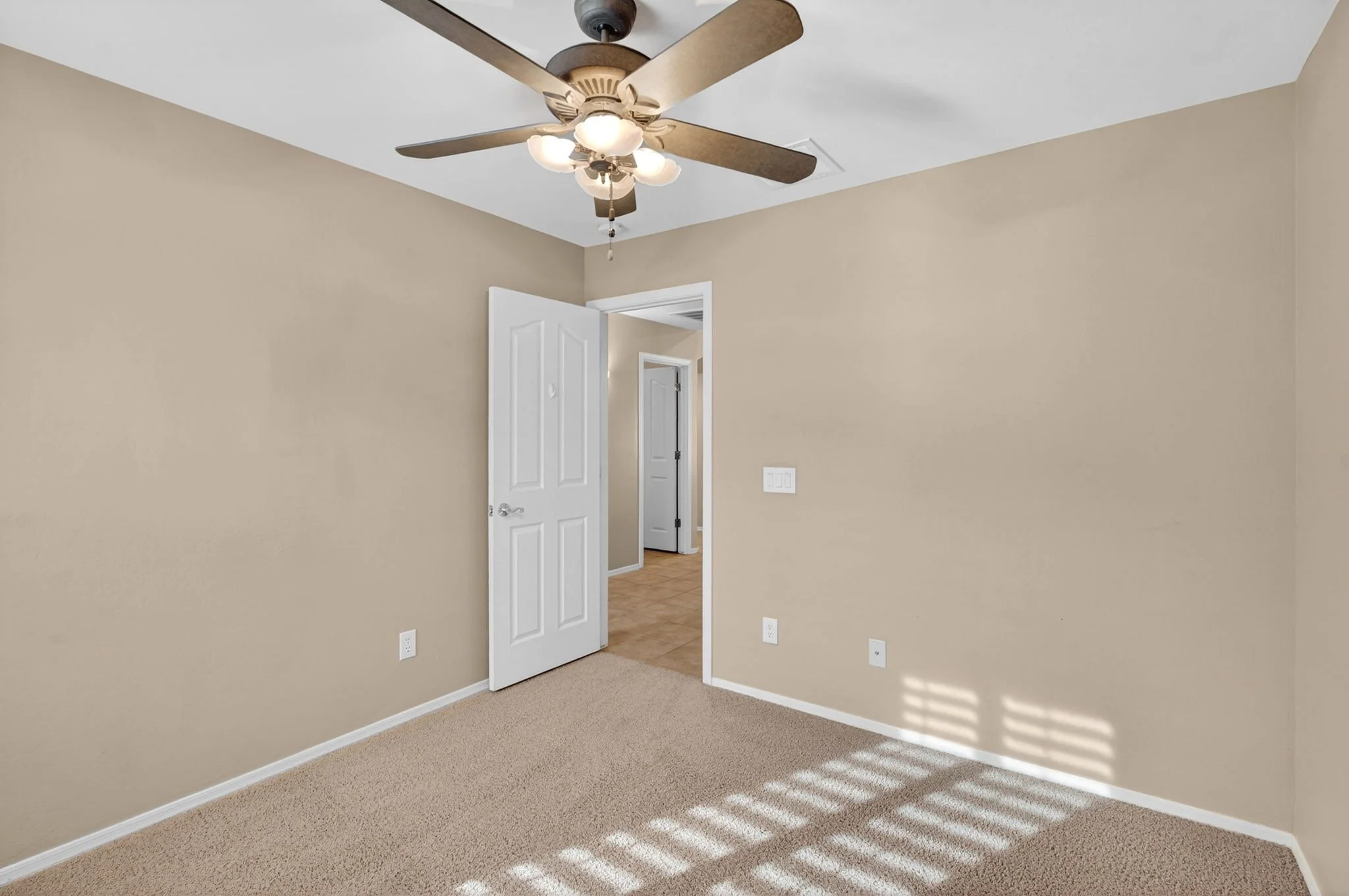 An empty beige room with a ceiling fan and light, open door leading to a hallway with tiled floor, sunlight casting shadows on the carpet.