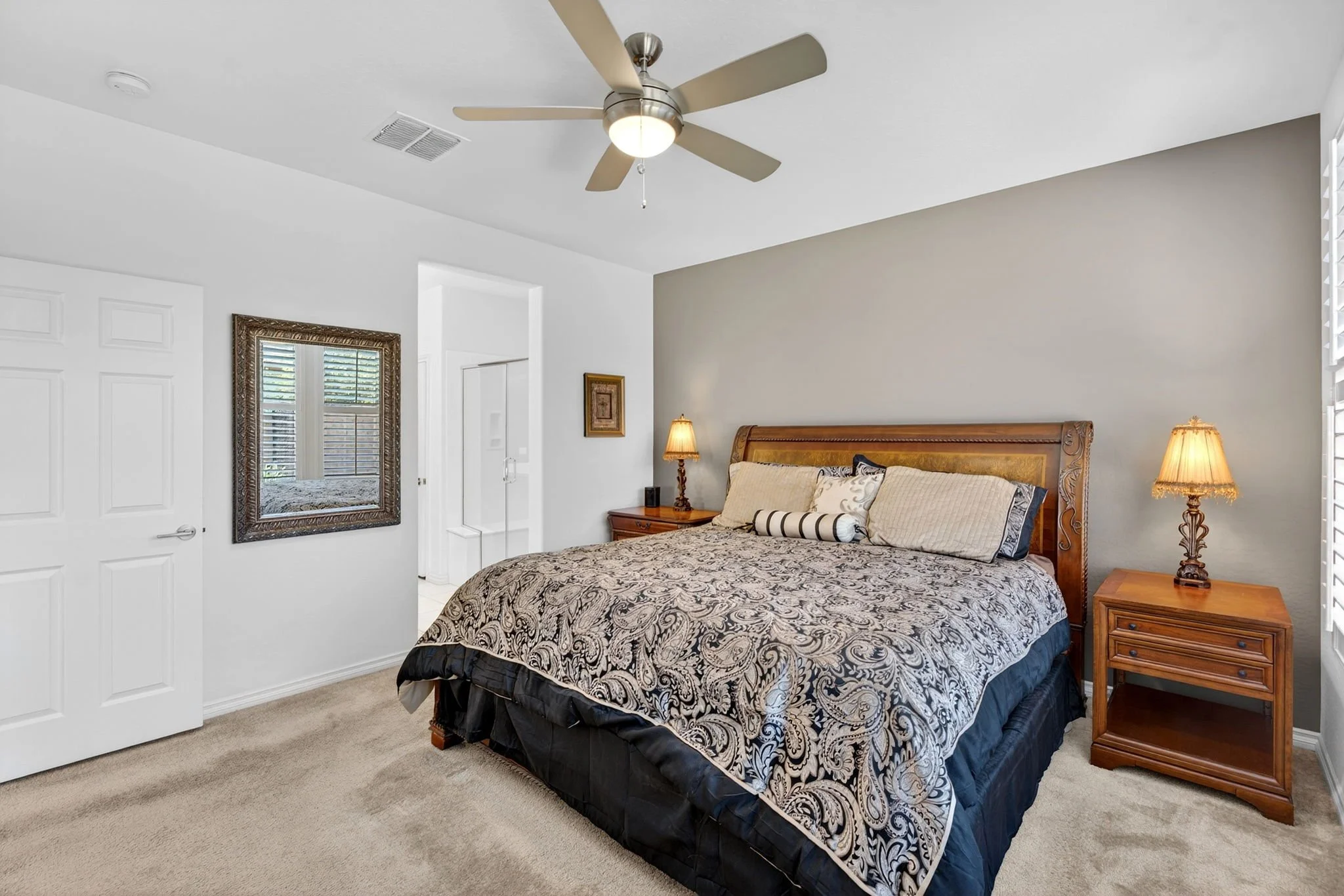 A bedroom with a wooden bed frame, patterned bedding, two nightstands with lamps, a wall mirror, and a ceiling fan with a light fixture.