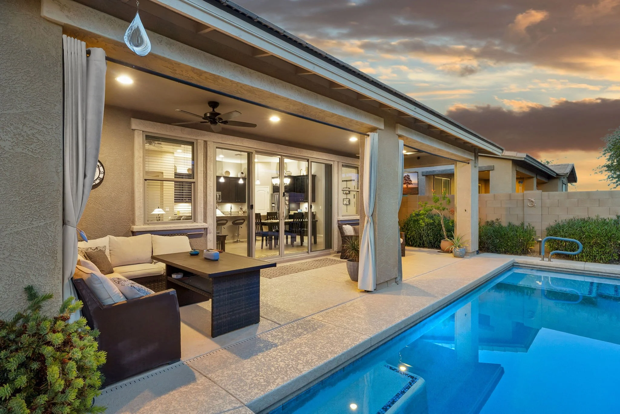 A backyard patio area at sunset featuring a swimming pool, outdoor seating with cushions, a dining table with chairs, sliding glass doors leading into the house, a ceiling fan, and surrounding plants and shrubs.