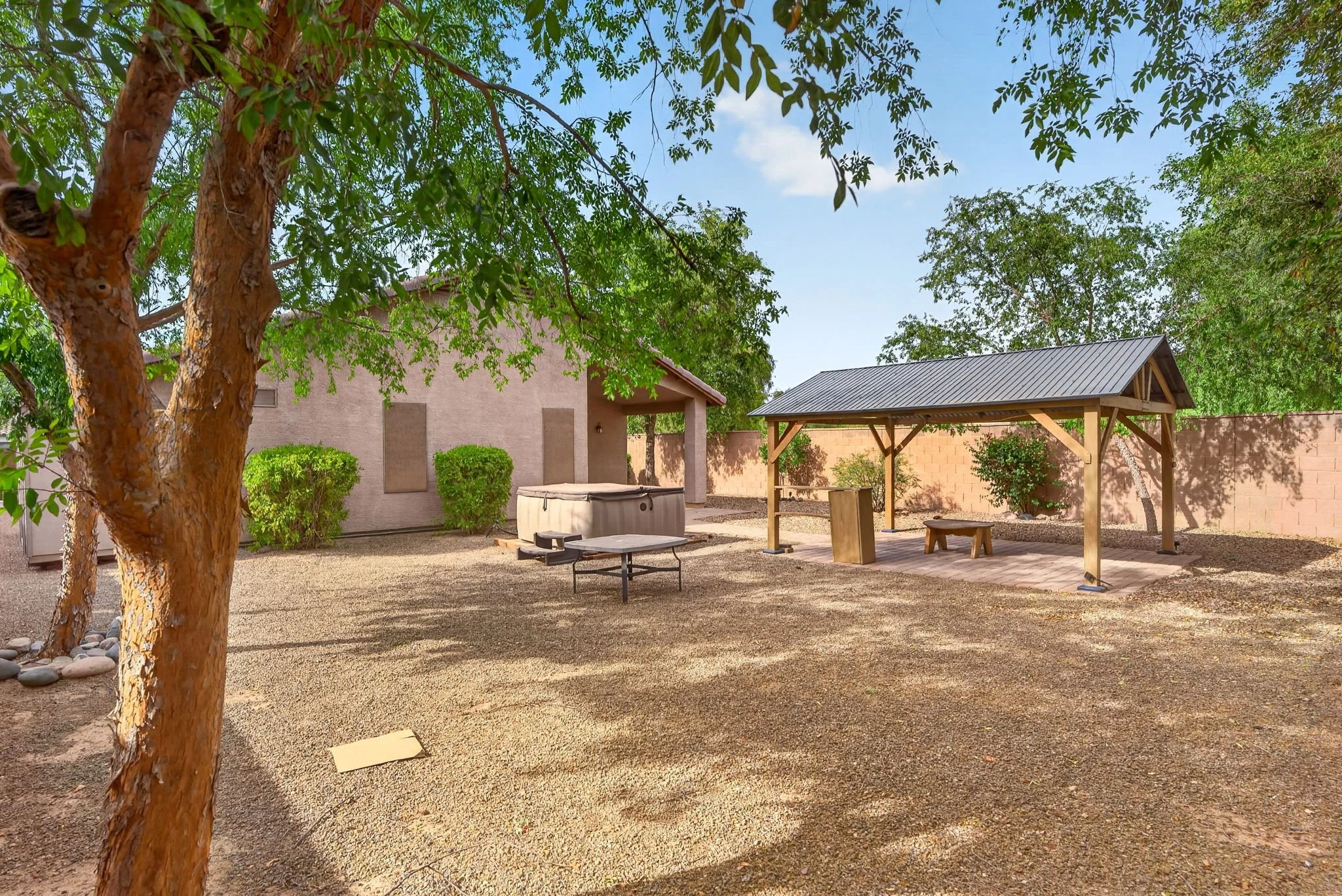 A backyard with a large tree in the foreground, a covered patio area, and a shed or small building with a hot tub nearby. The yard is enclosed by a beige block wall.
