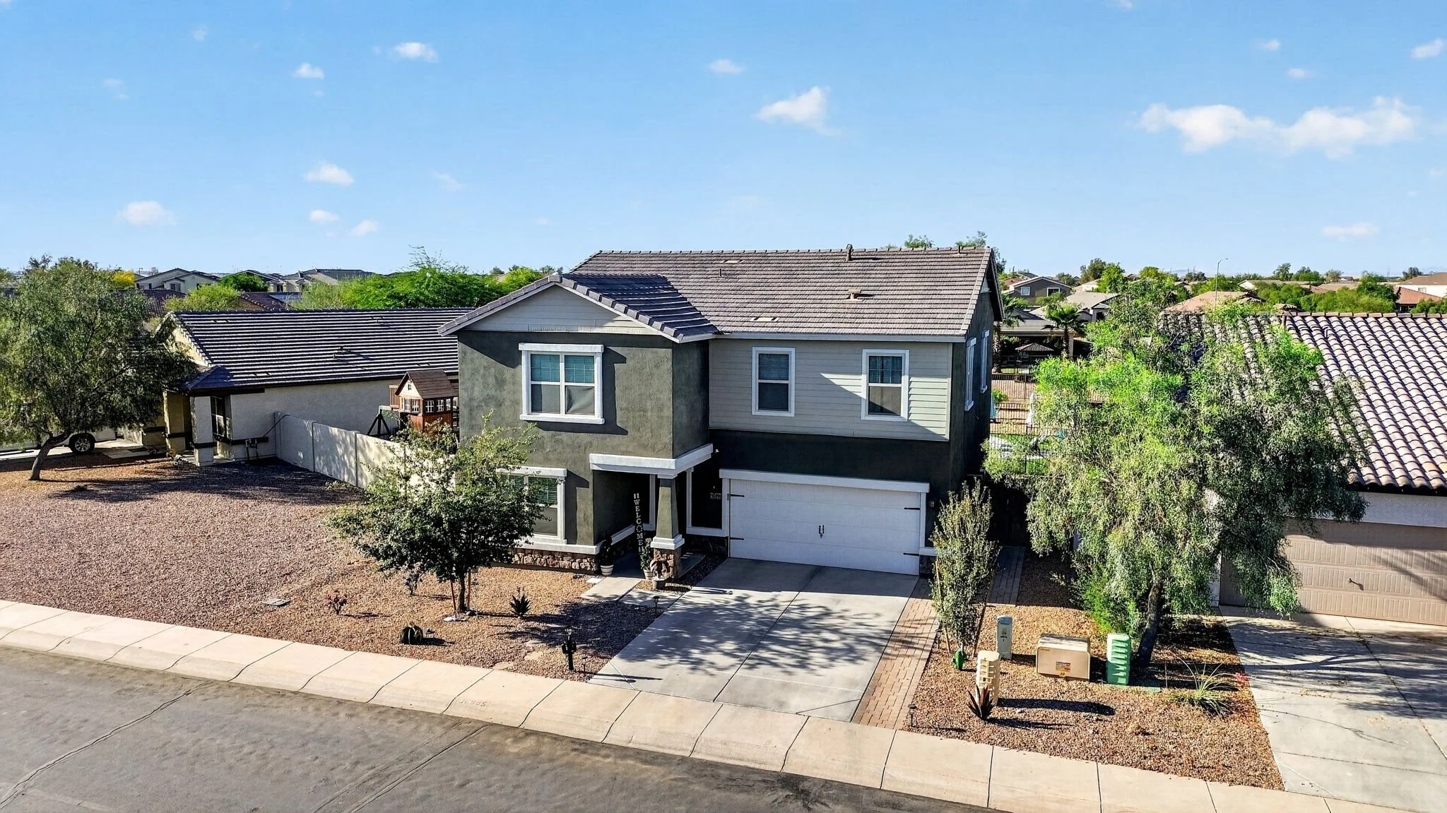 A two-story suburban house with a front porch, white garage door, and a landscaped front yard with small trees and desert plants, under a clear blue sky.