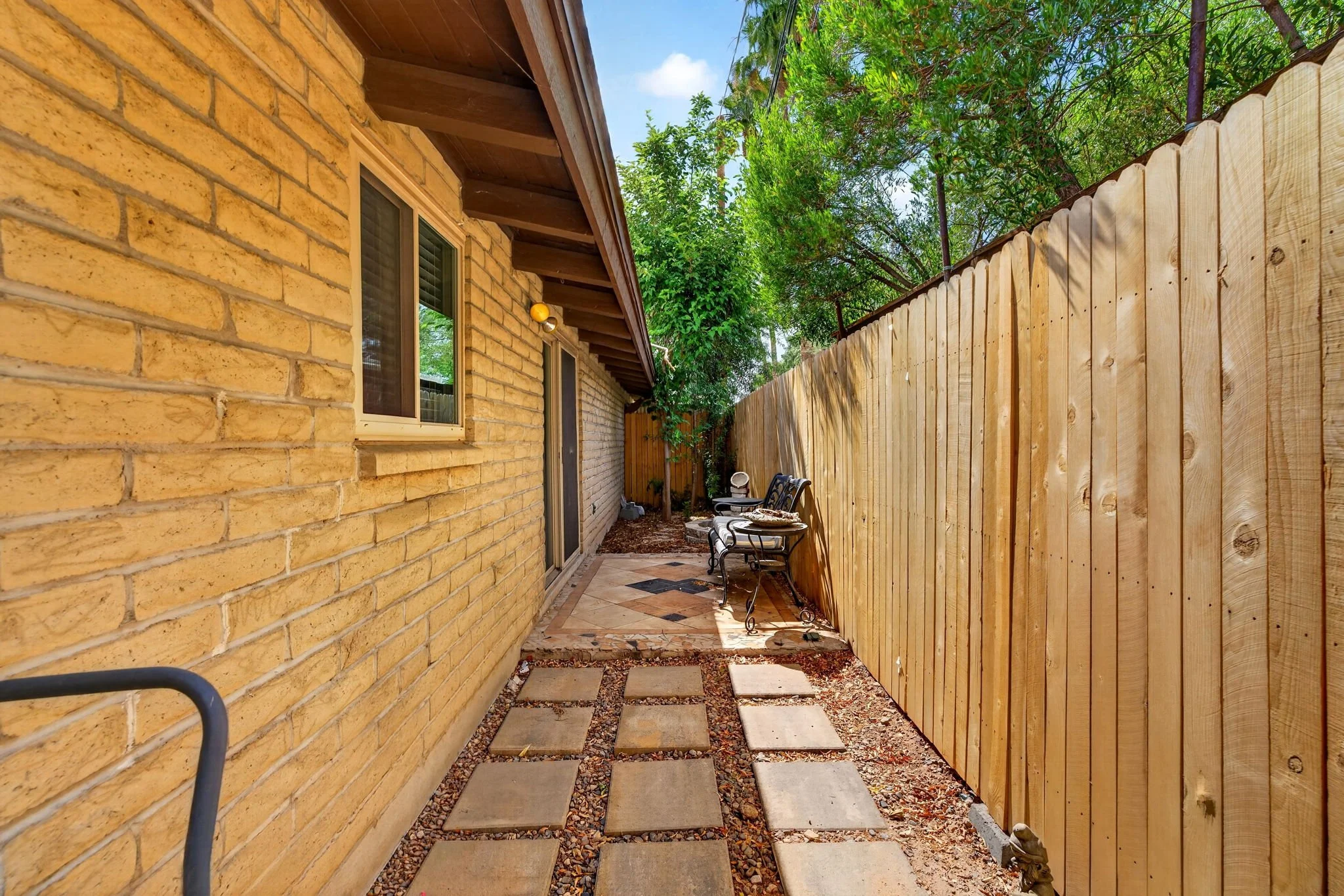 A small backyard patio area with a brick house on the left, a dirt and gravel ground with large paver stepping stones, and a tall wooden fence on the right. There are two metal chairs and a small table in the corner near the house. Behind the fence, 