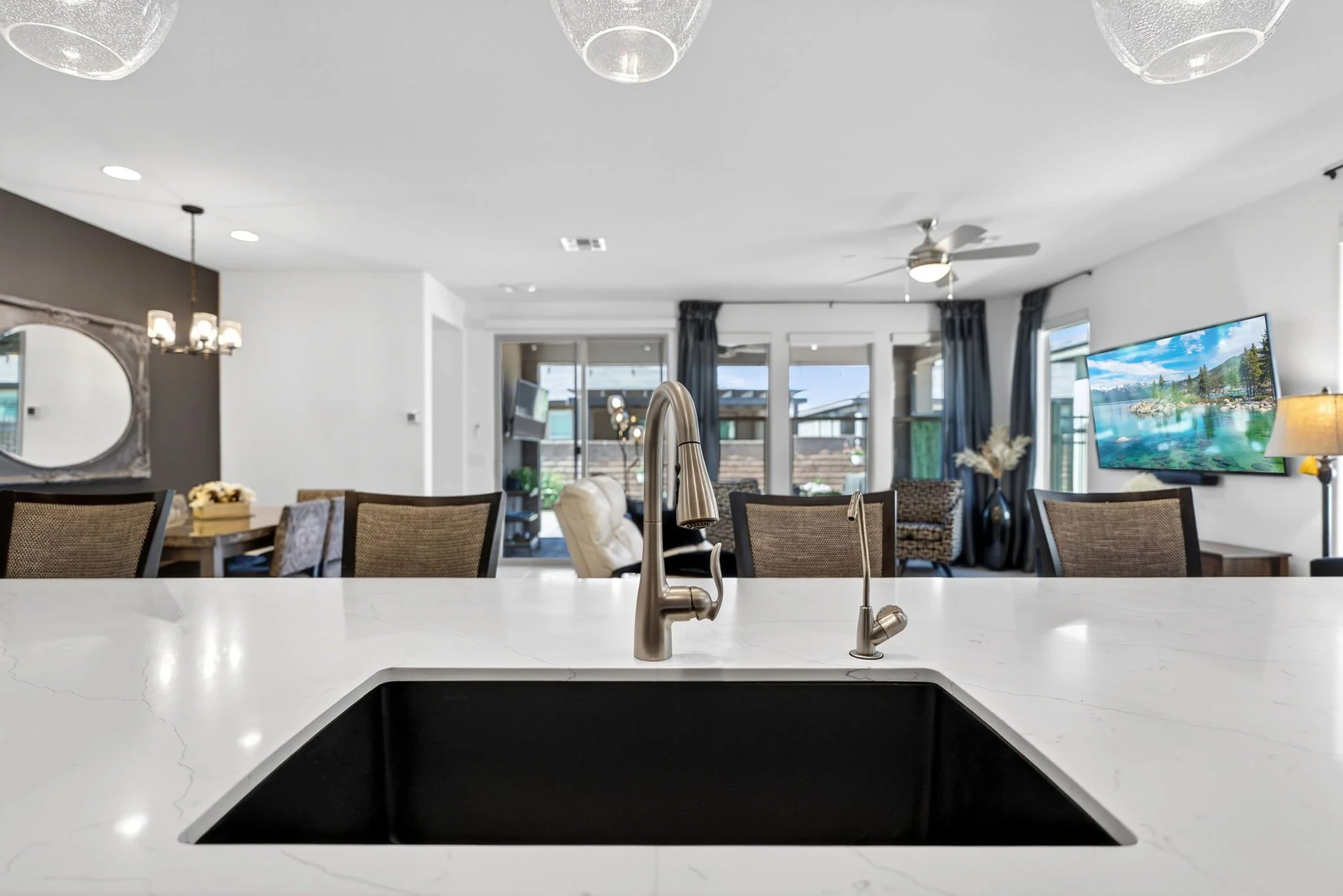 Modern kitchen with white marble island and stainless steel sink, overlooking a living room with large windows, a television, and outdoor patio.