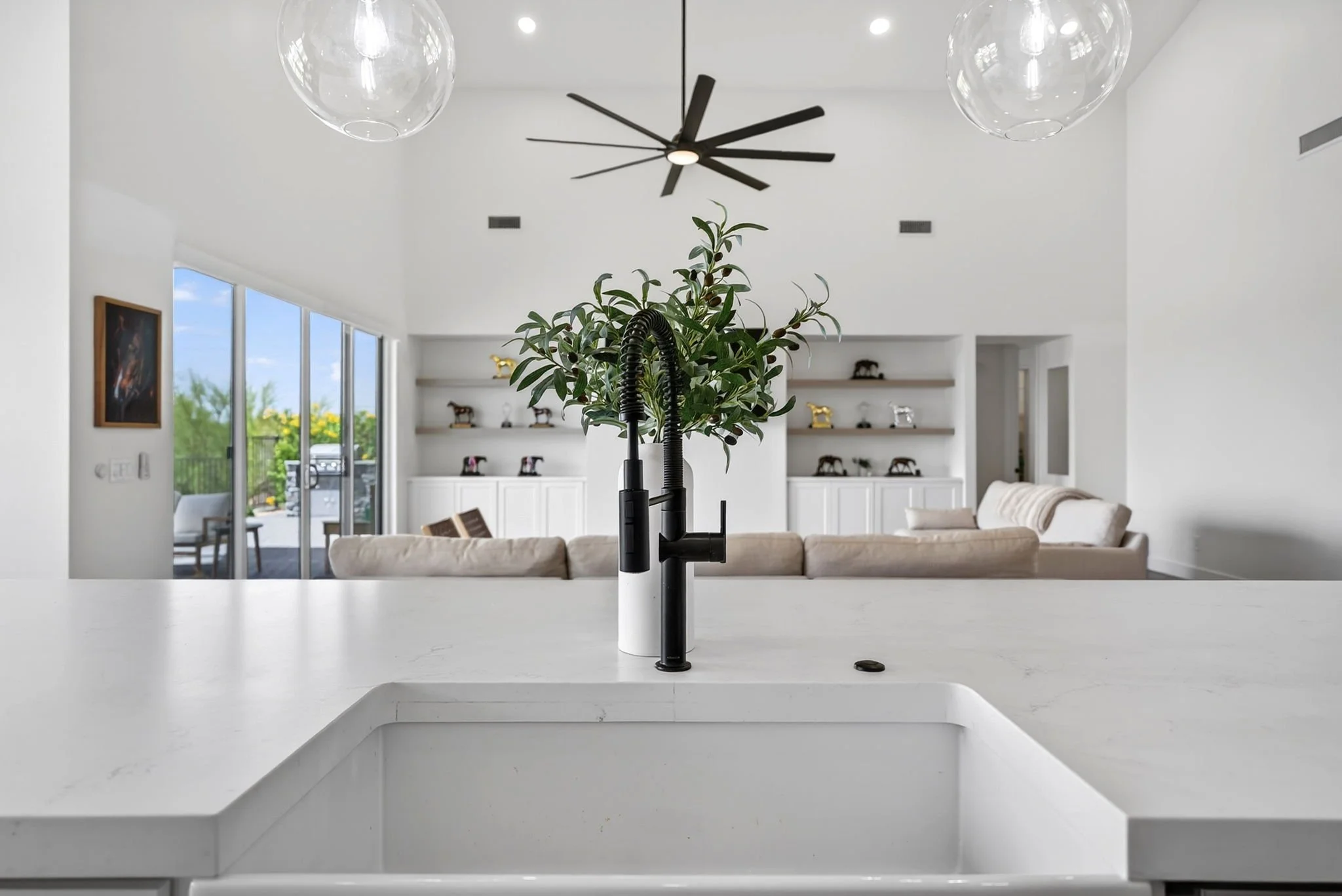 Modern kitchen with white countertops and a black faucet, a large green plant in the sink, and a living room with white sofas and built-in shelves with decorative items in the background.