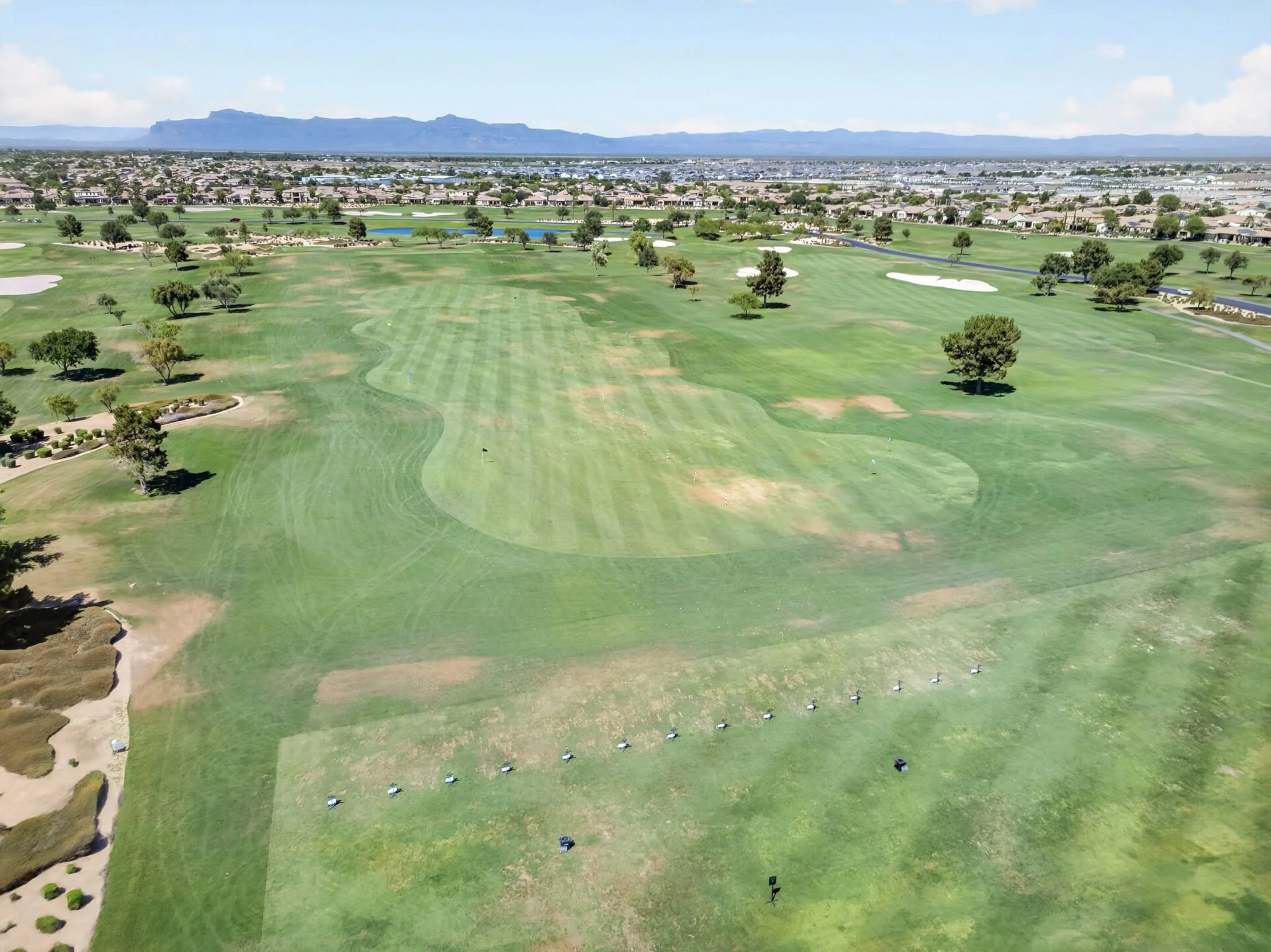 Aerial view of a golf course with green fairways, sand traps, trees, water hazards, and holes, with urban development and mountains in the background.
