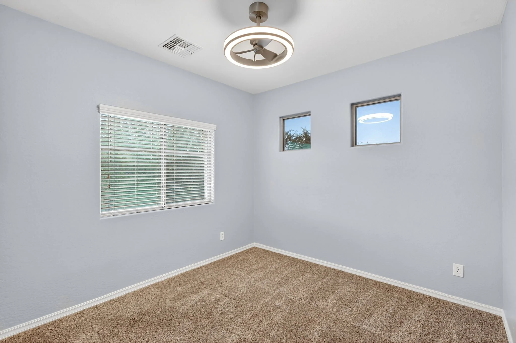 Empty room with light blue walls, a window with white blinds, and two small windows near the ceiling. There is a circular ceiling fan with a light, beige carpeting, and a white electrical outlet on the wall.