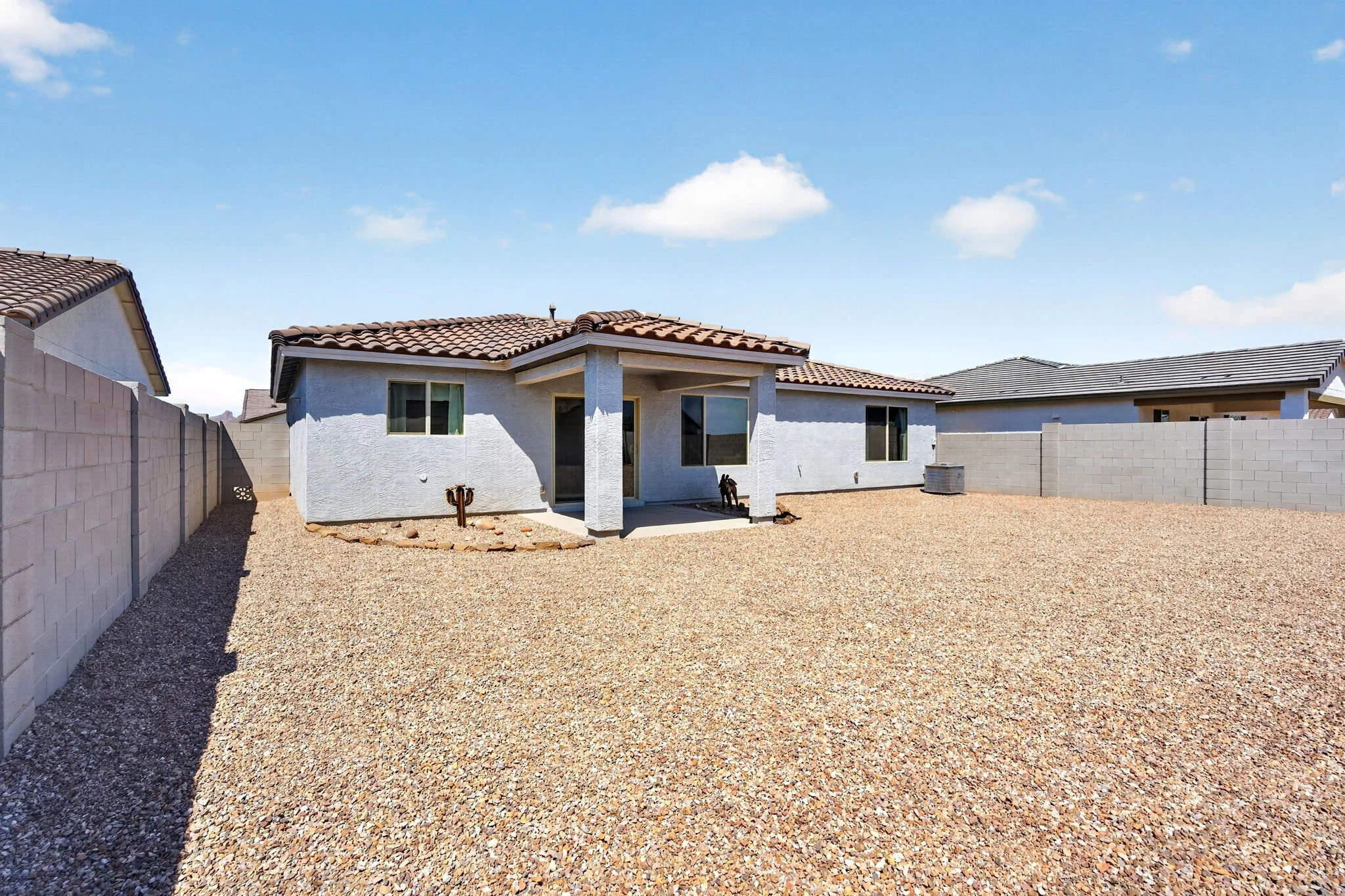 Backyard with gravel ground, white house with tile roof, surrounded by a concrete block wall, under a blue sky with a few clouds.