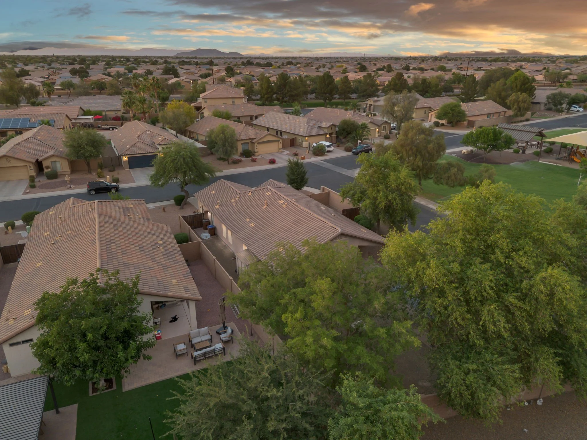 An aerial view of a suburban neighborhood during sunset, showing houses with tiled roofs, trees, lawns, and a playground in the park.