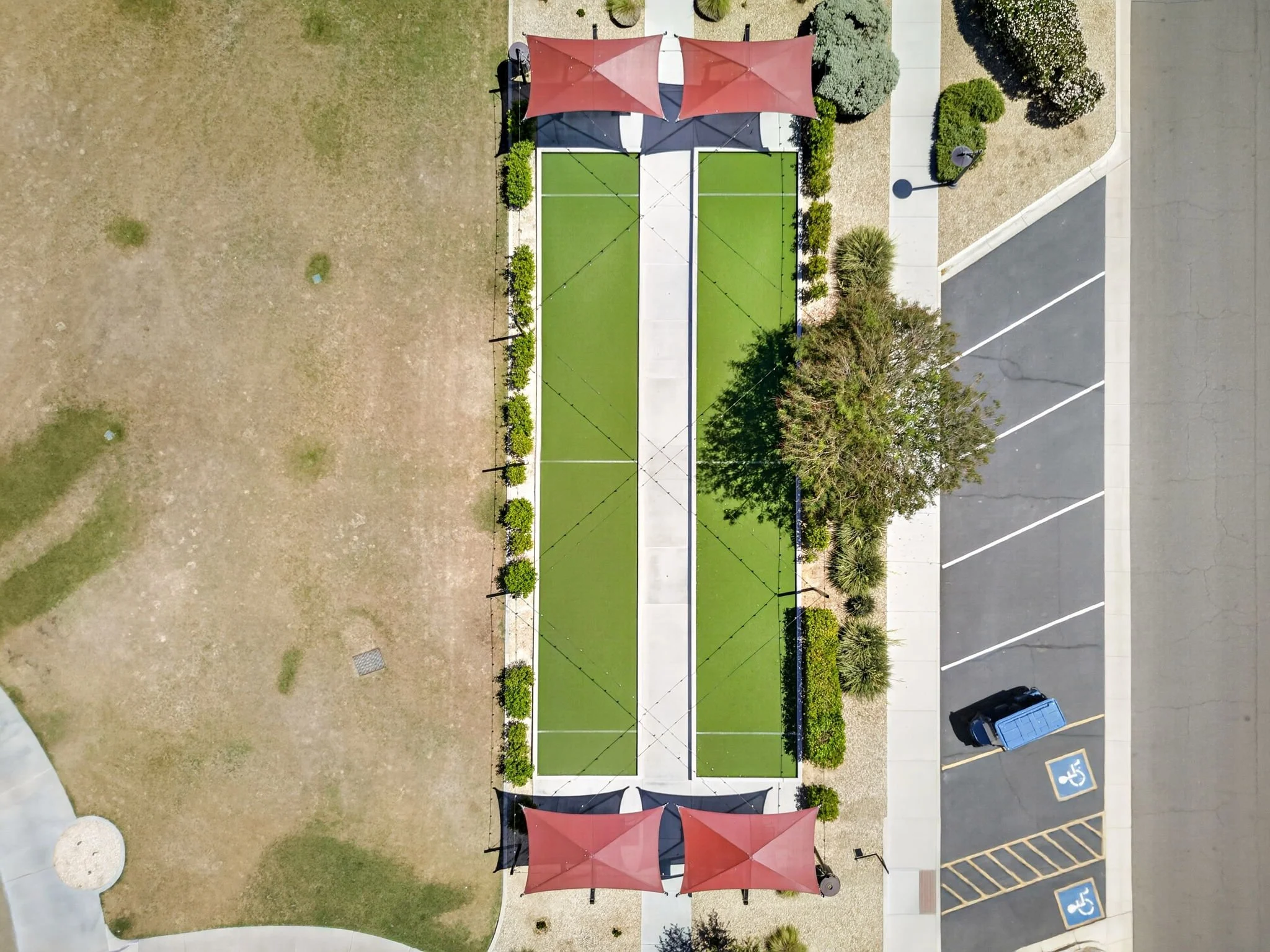 An aerial view of a small outdoor sports court with two shaded seating areas and surrounding landscaped bushes, trees, and sidewalks. There is a parking lot with a blue trash bin nearby.