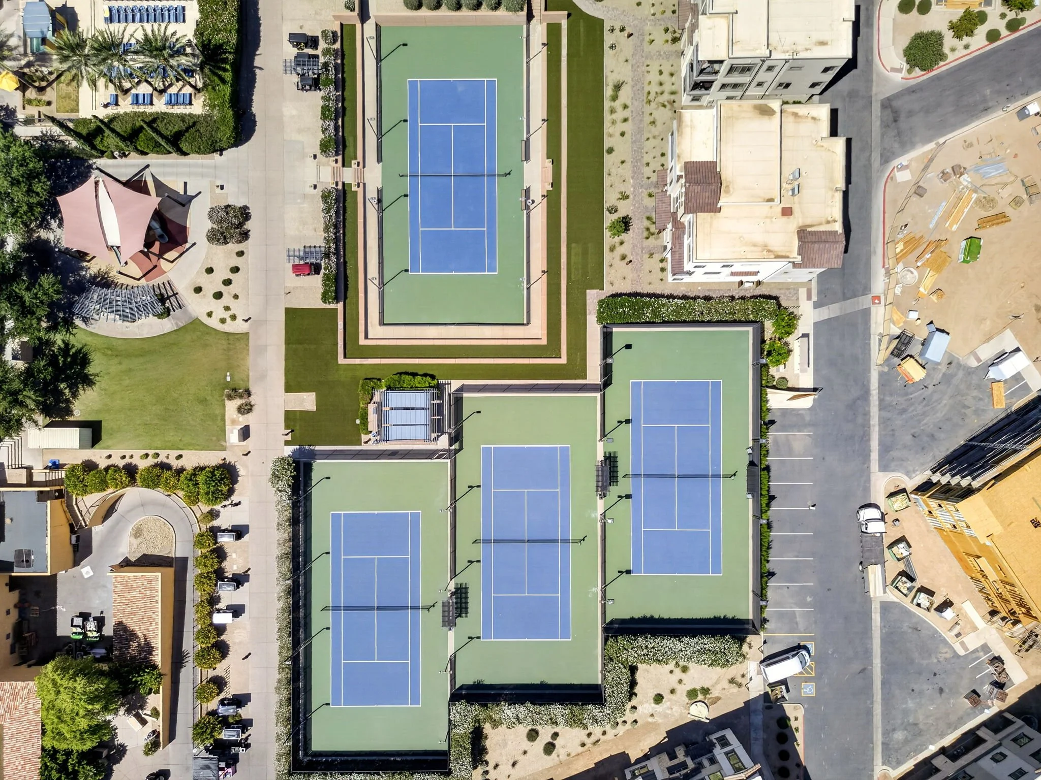 Aerial view of a sports complex featuring multiple tennis courts with blue surfaces and surrounding green areas, adjacent to a construction site with building materials and vehicles.
