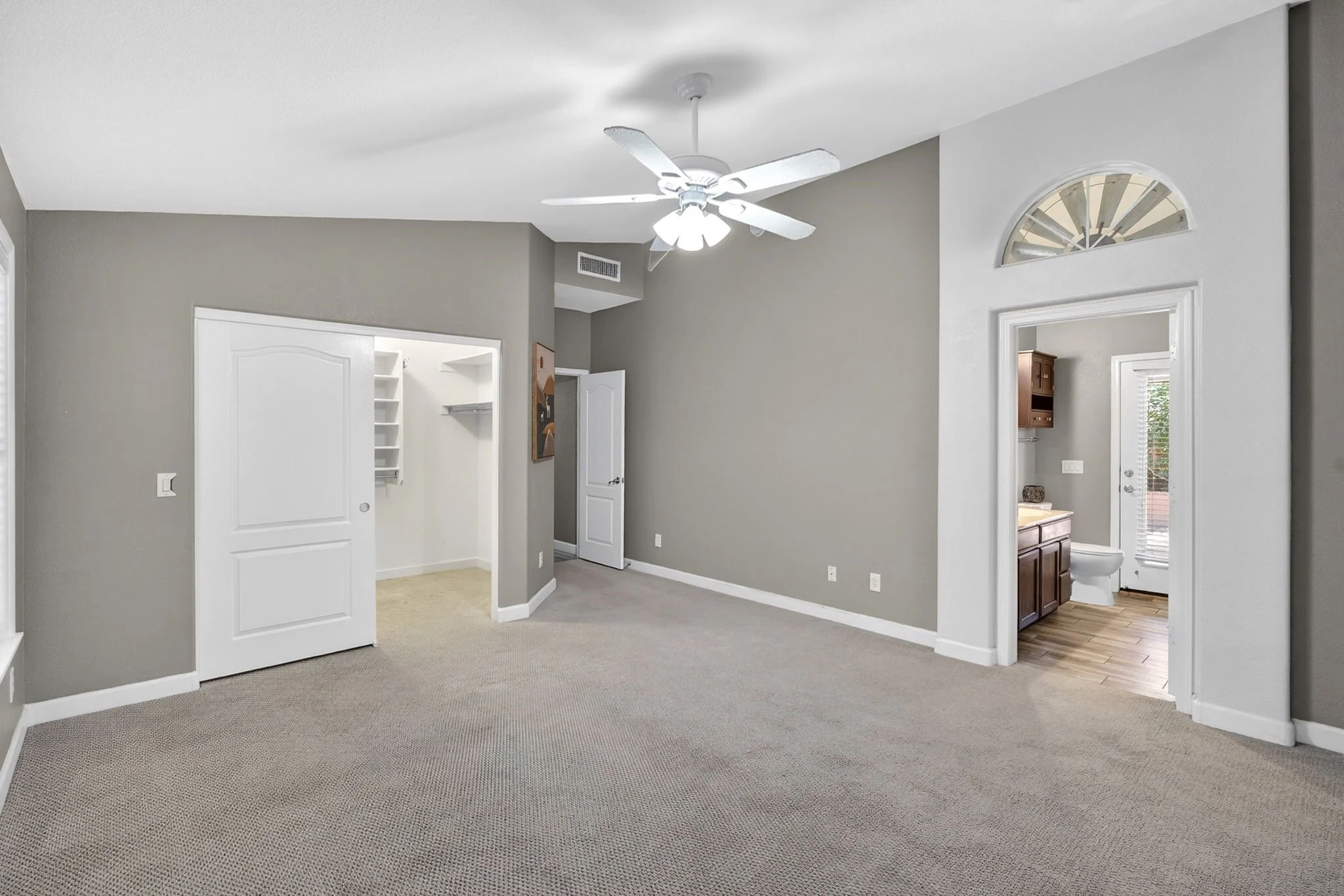 Empty living room with gray walls, beige carpet, and a white ceiling fan. There are doorways leading to a bathroom and another room, with sunlight coming through a window.