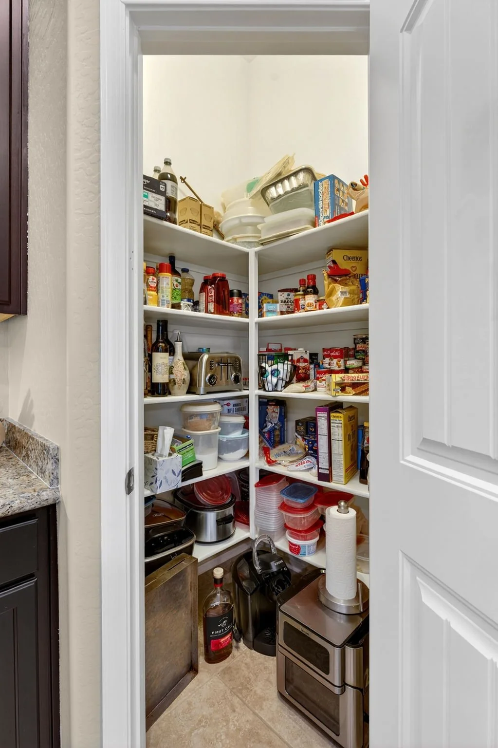 Pantry shelves filled with food, condiments, kitchen appliances, and containers, with a paper towel roll on a stand on the floor.