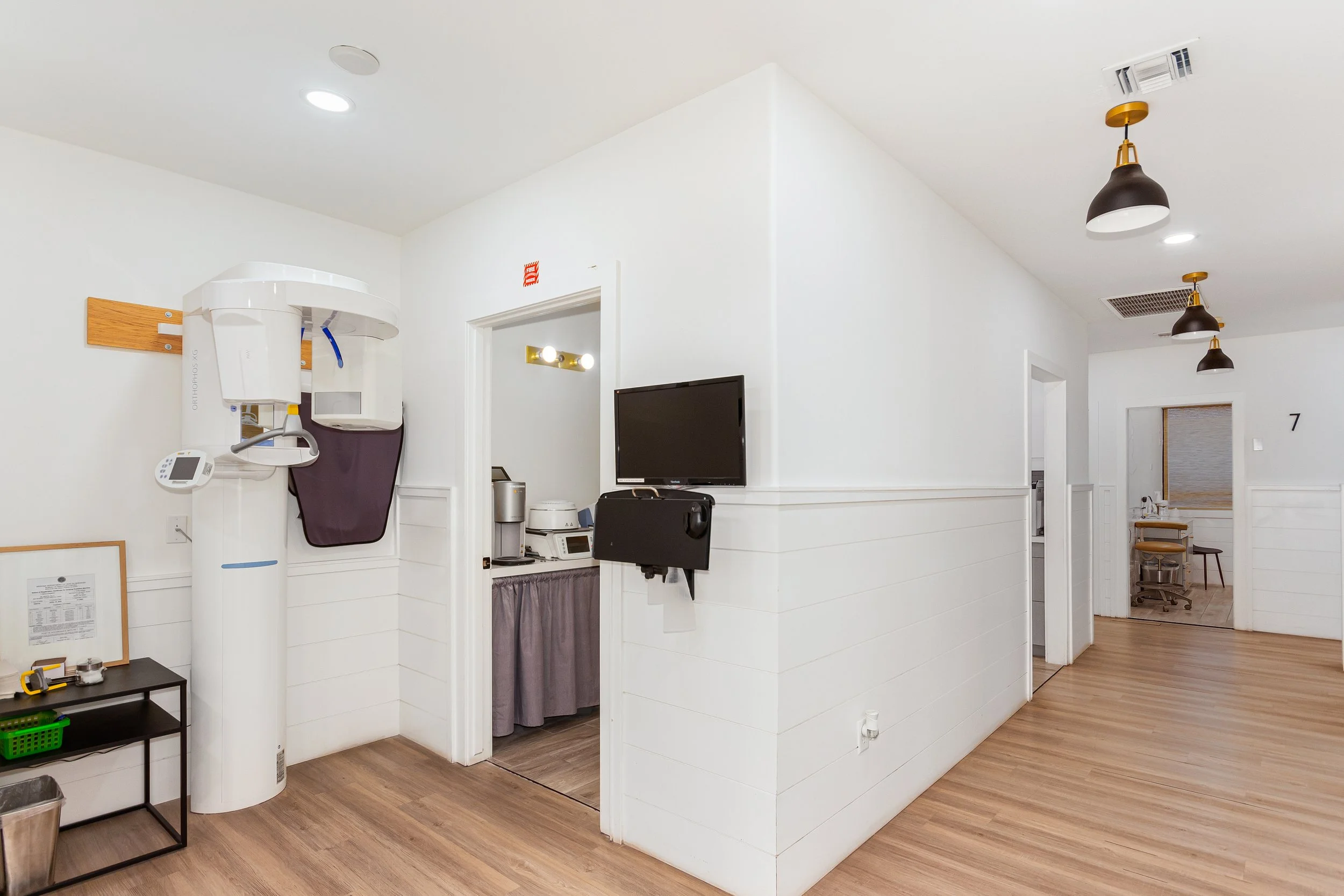 Interior view of a medical or office space with white walls, wood flooring, and ceiling lights. Medical imaging equipment is visible, including a panoramic X-ray machine. There are doors, a TV screen, and various small furniture pieces.
