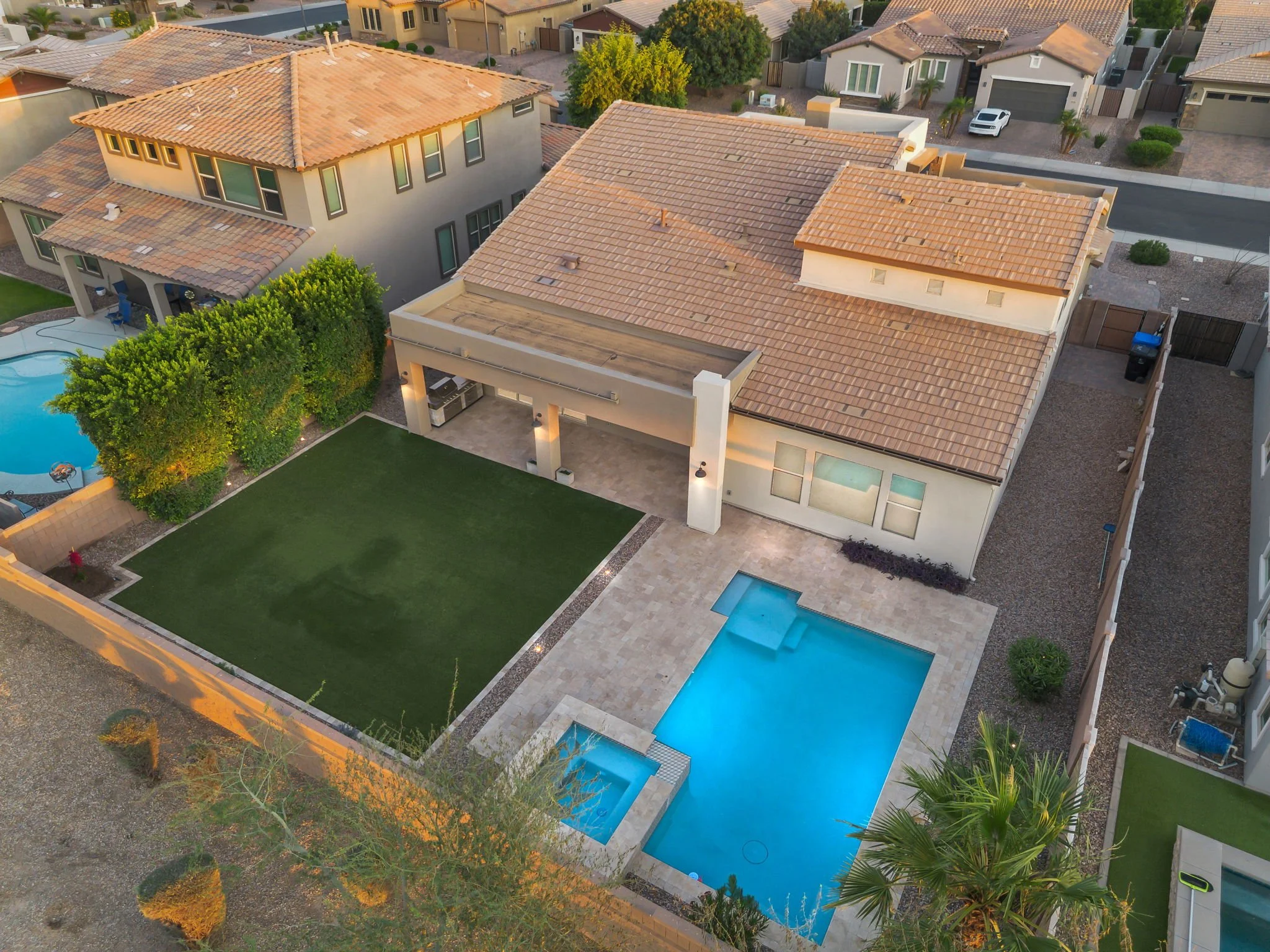 Aerial view of a residential backyard with a swimming pool, a well-maintained lawn, trees, and neighboring houses with tiled roofs.