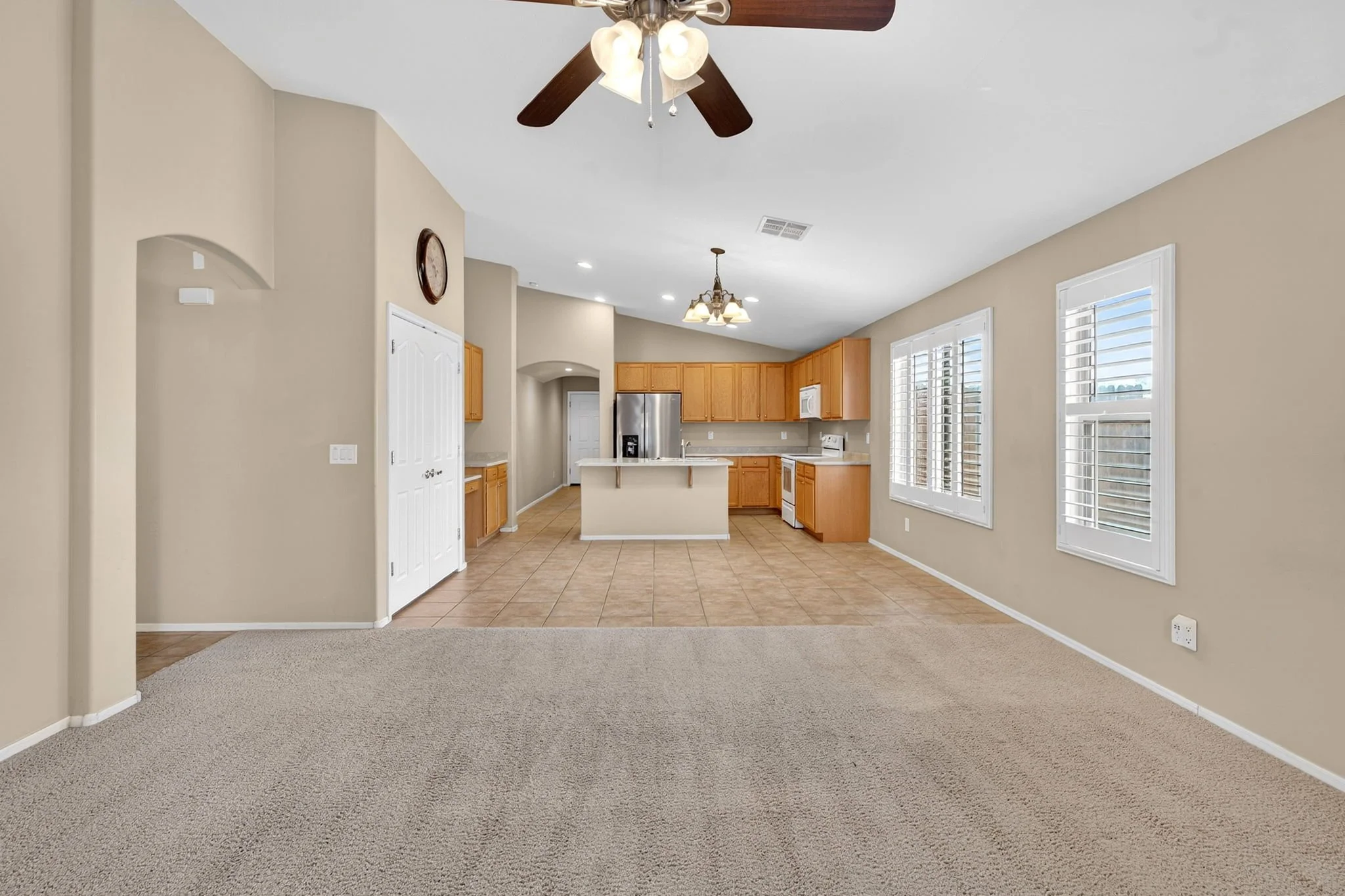 Open kitchen and living area with beige walls, large windows with blinds, tiled floor in kitchen, carpeted floor in living area, wooden kitchen cabinets, white appliances, ceiling fans with lights.