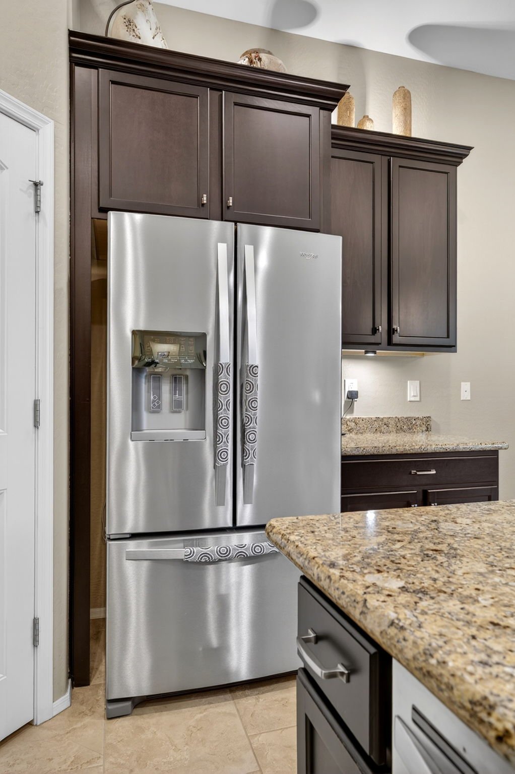 Modern kitchen with stainless steel Whirlpool refrigerator, dark wood cabinets, granite countertops, and beige tiled floor.