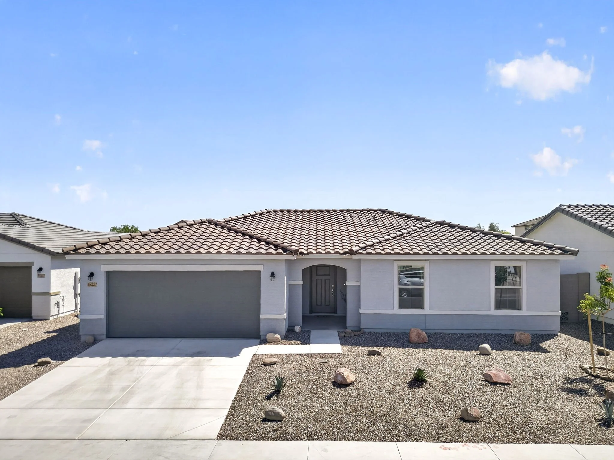 Front view of a modern house with a grey garage door, dark front door, tiled roof, and a landscaped yard with rocks and small plants under a clear blue sky.
