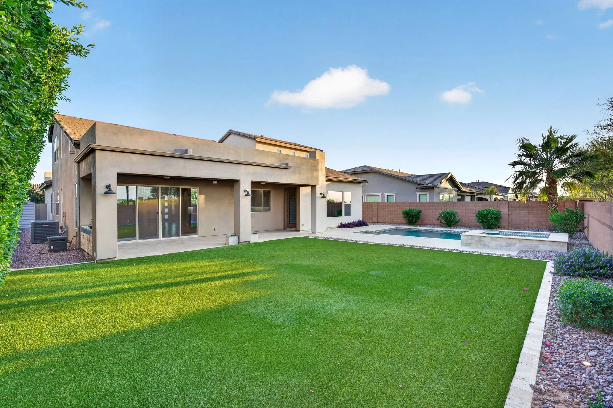 A backyard with green grass, a swimming pool, a modern house with a patio, and a clear blue sky with a few clouds.