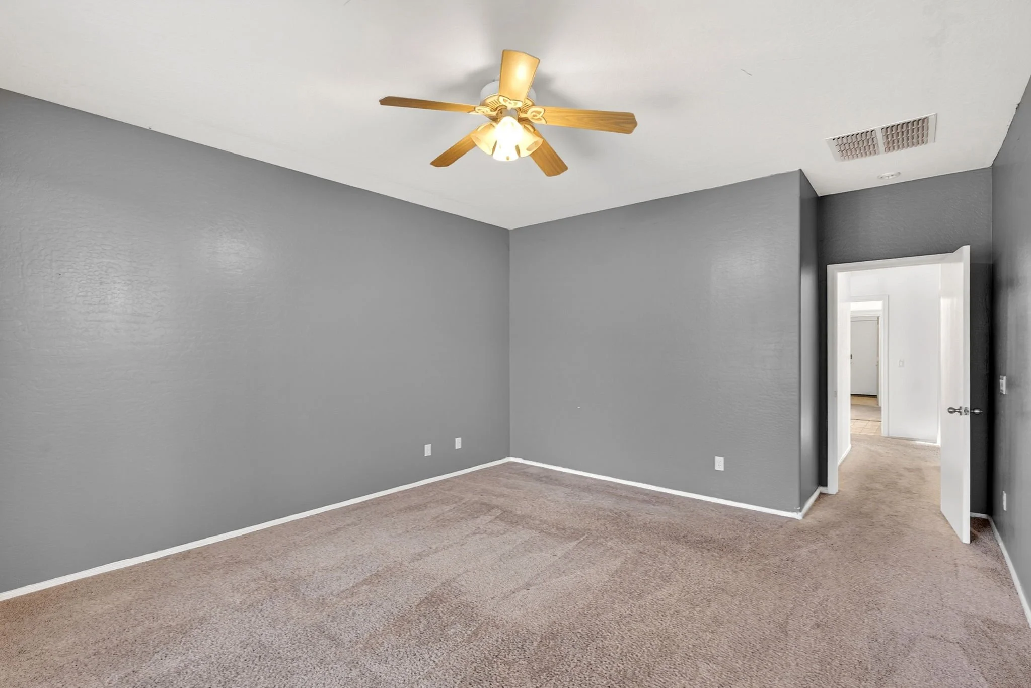 Empty room with gray walls, a ceiling fan with lights, a doorway leading to another room, and beige carpet flooring.