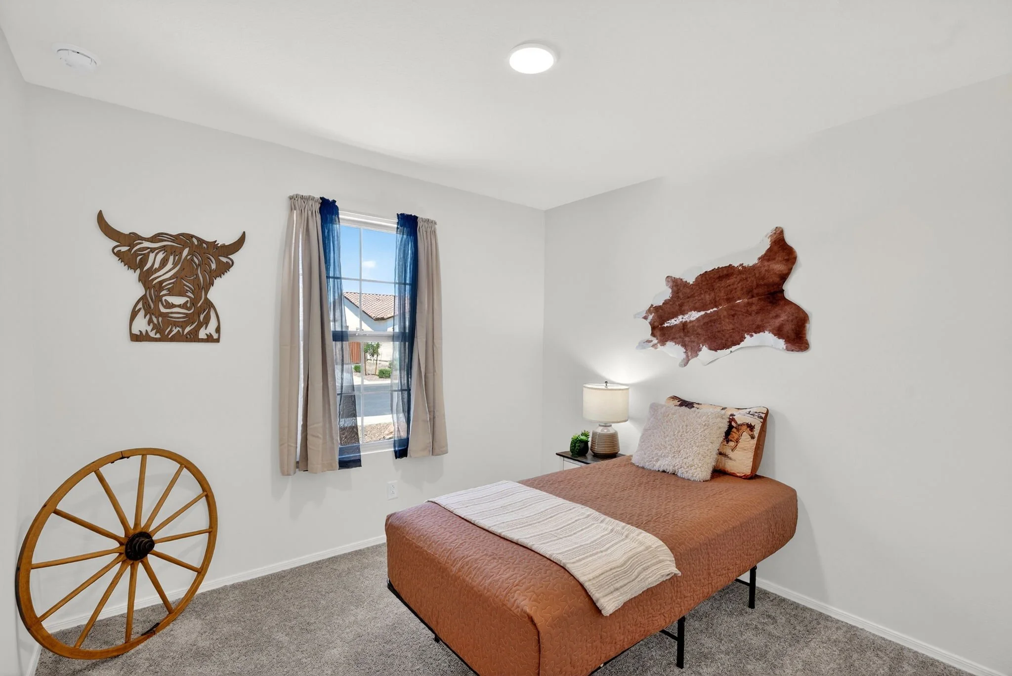 A bedroom with a bed, a nightstand with a lamp, wall decor including a wooden highland cow head and a red map outline, a window with blue and beige curtains, and part of a wooden chair.