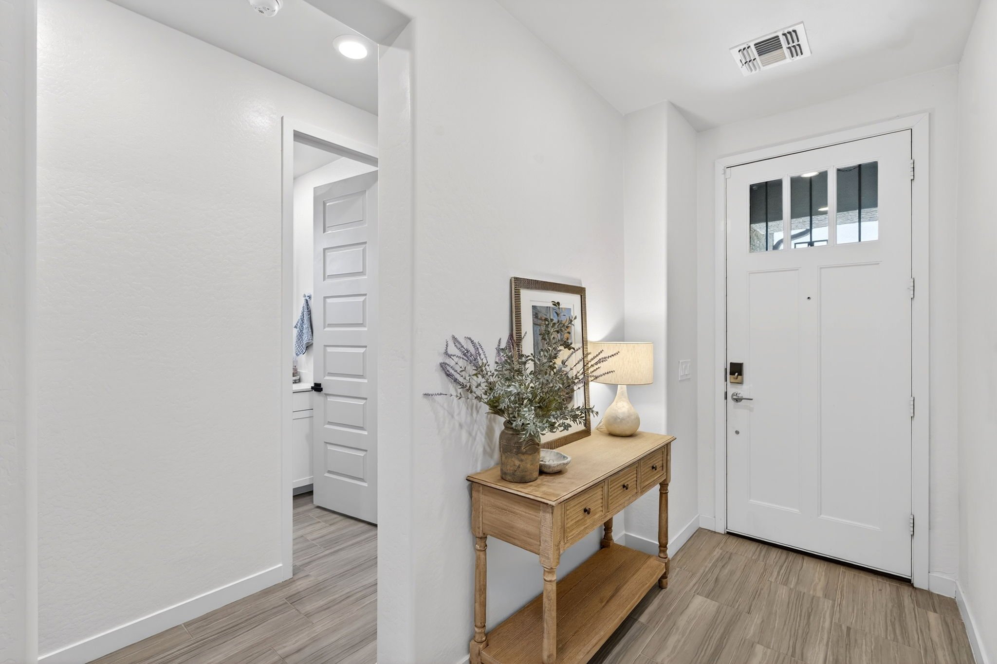 Entryway with white walls and door, wooden console table with a potted plant and framed picture, beige rug, and ceiling air vent.