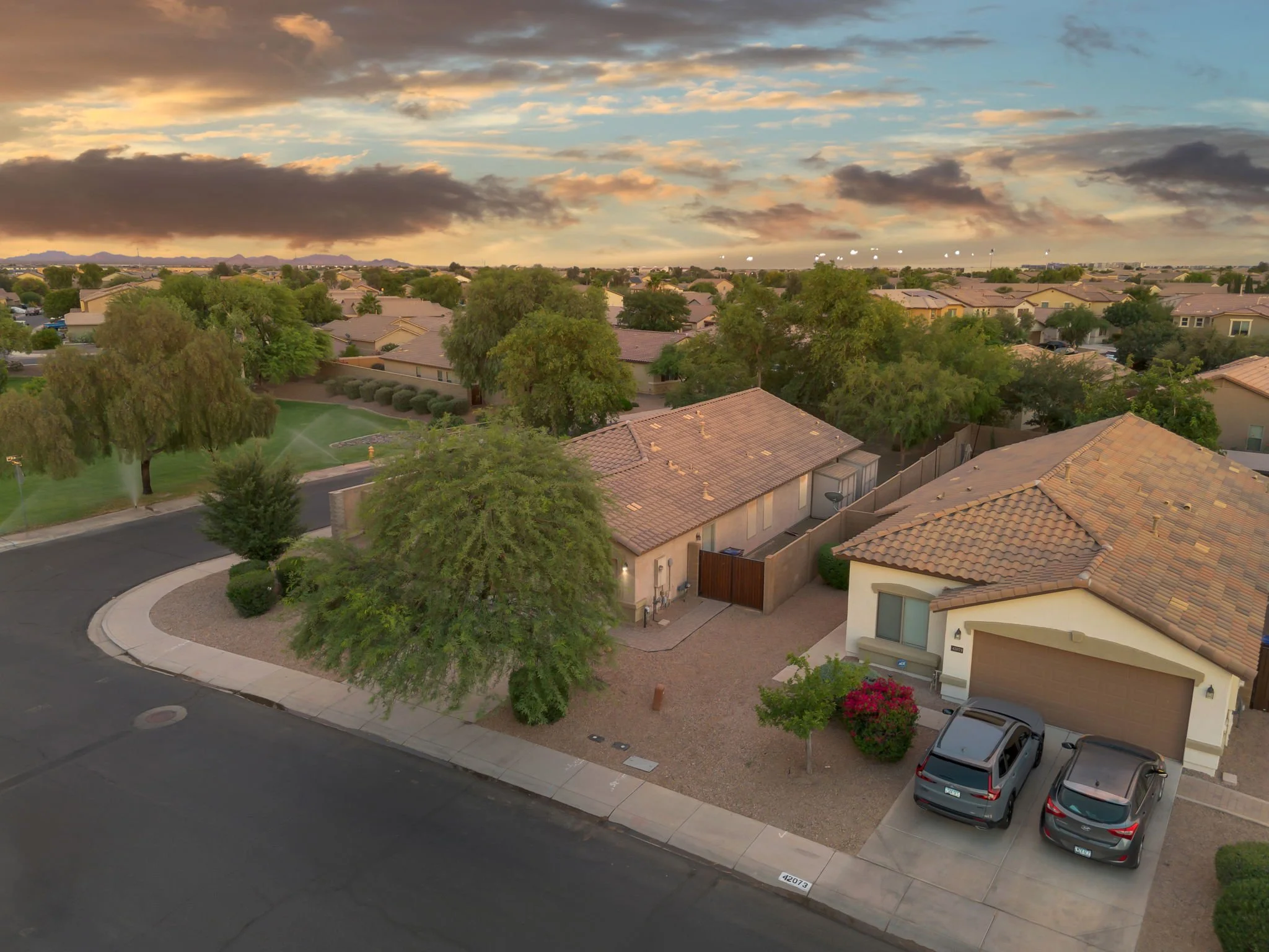 A suburban neighborhood scene at sunset with houses, trees, parked cars, a golf course, and a cloudy sky with patches of blue.