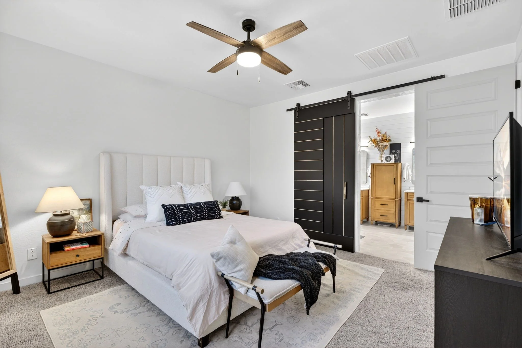 A bedroom with a white bed, wooden nightstand, and beige lamp. A black blanket is at the foot of the bed. A sliding barn door leads to a room with a wooden dresser and flowers. A flat-screen TV is on a black stand. The ceiling has a wooden fan with t