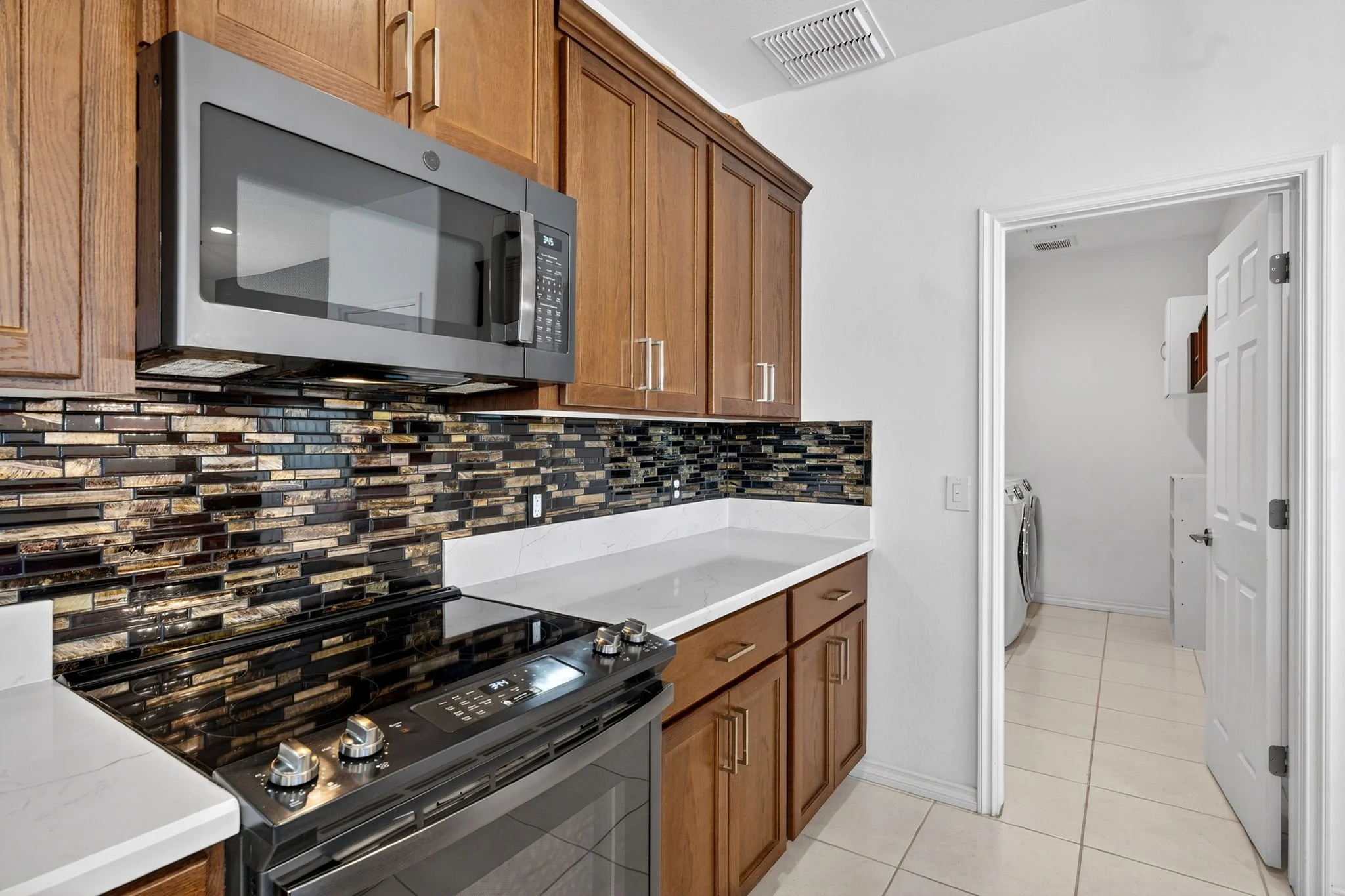 Kitchen with wooden cabinets, a stainless steel microwave, a stove, and a laundry area with a washing machine visible through an open door.