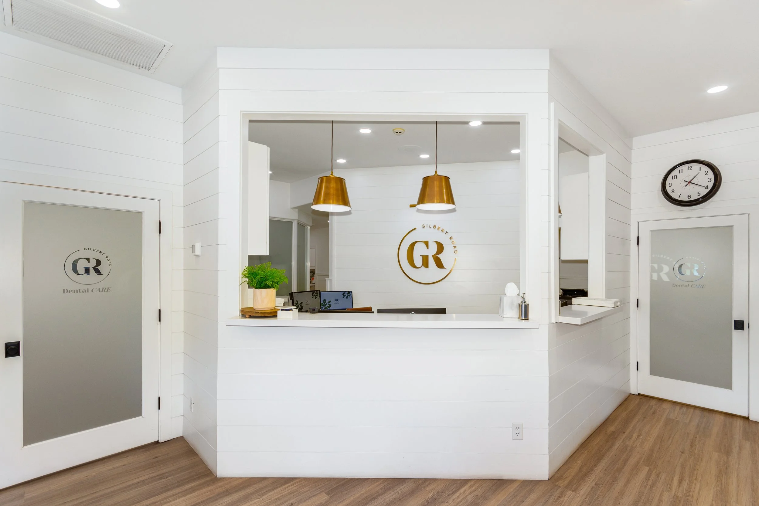 Reception area of a dental clinic with white walls, a reception desk, two large gold pendant lights, a clock on the wall, a plant on the desk, and a logo that reads 'Gilbert Road Dental Care'.