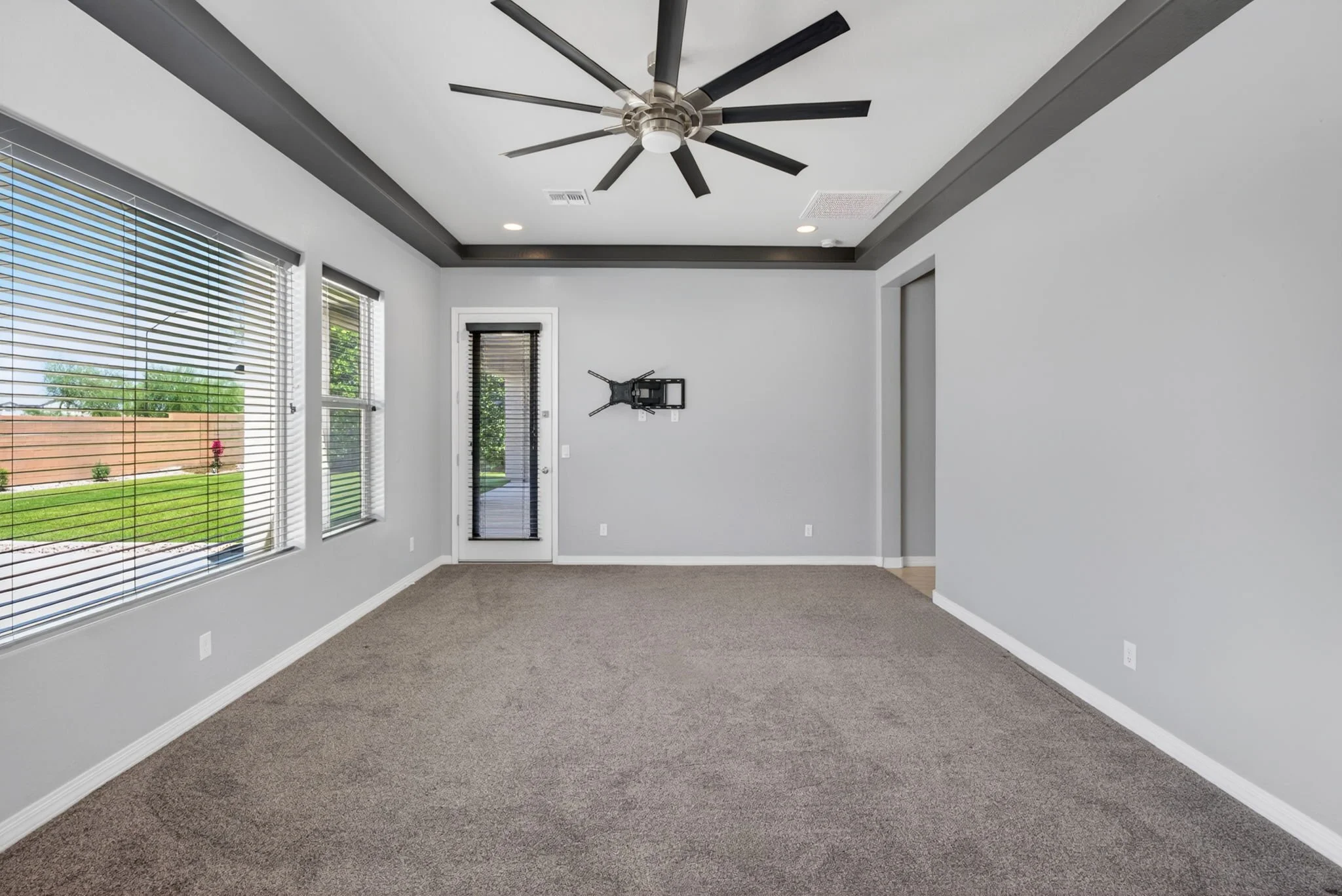 Empty living room with gray walls, large windows with blinds, a carpeted floor, a ceiling fan, and a door leading outside.
