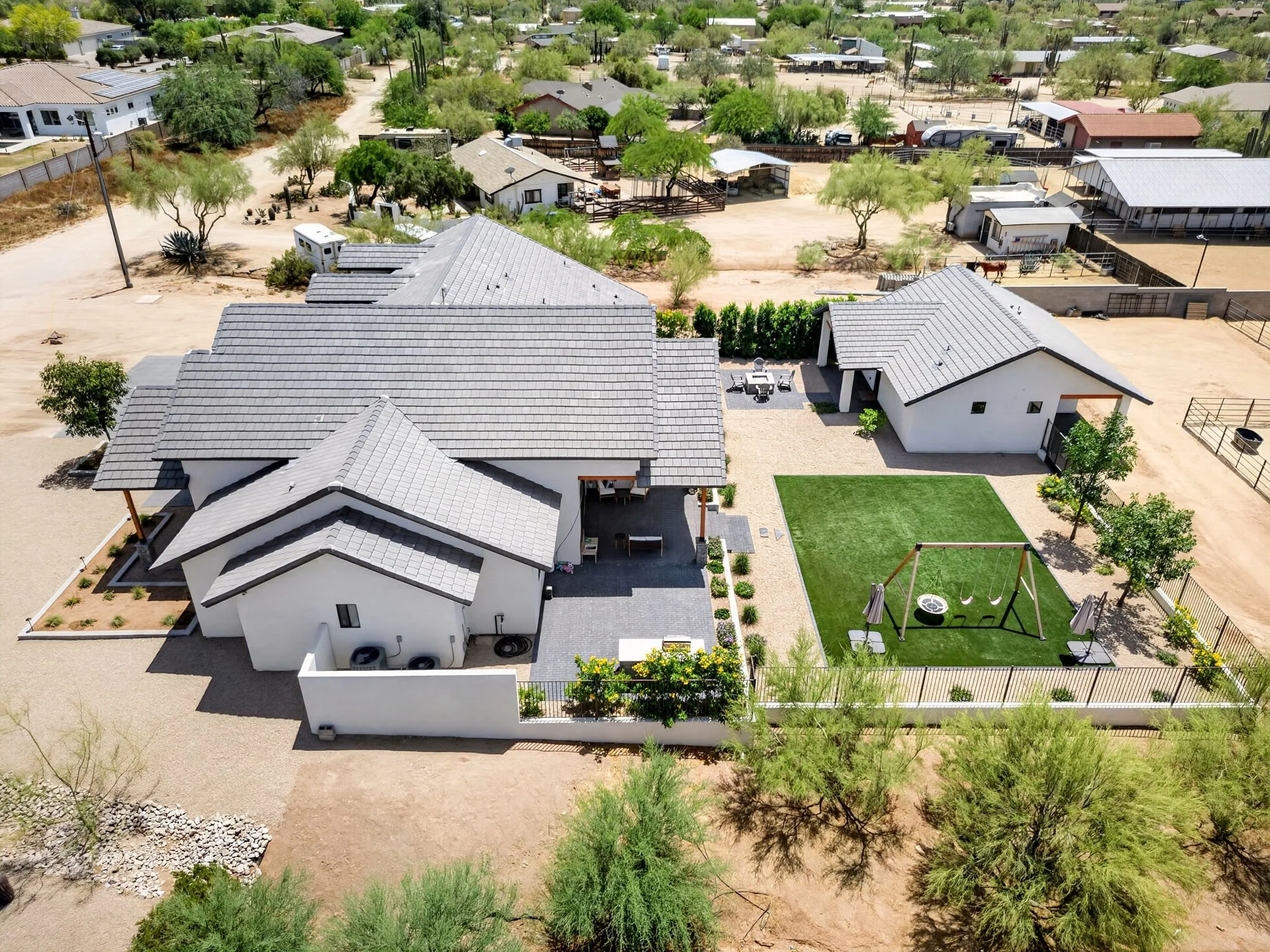 An aerial view of a residential backyard featuring a house with a grey tiled roof, a patio with outdoor furniture, a lush green lawn, and a fenced area with trees and plants, surrounded by other houses and desert landscape with cacti.