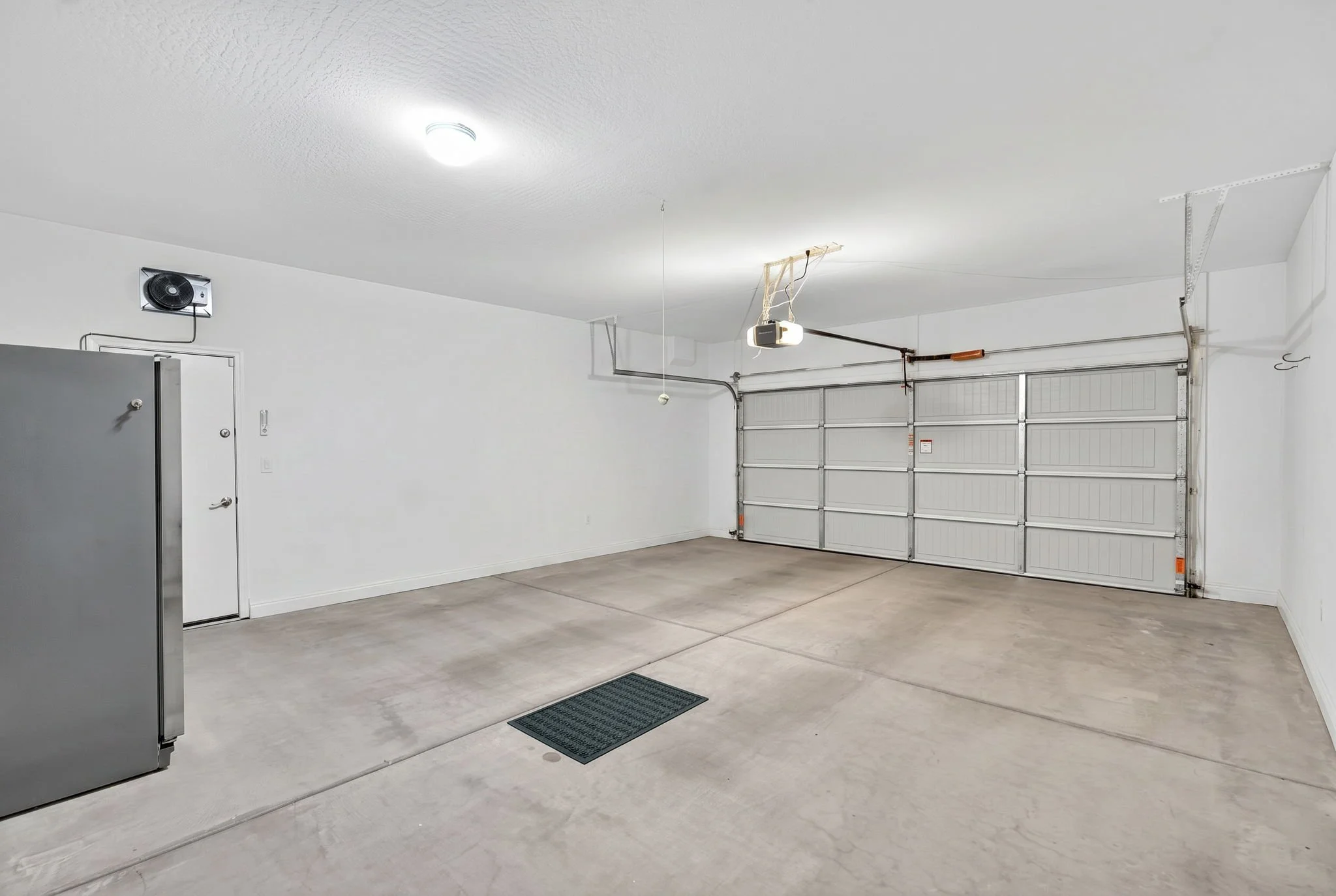 Empty garage with white walls, concrete floor, and a large garage door. There is a ceiling light, a ceiling-mounted garage door opener, a small utility cabinet, and a wall-mounted vent.