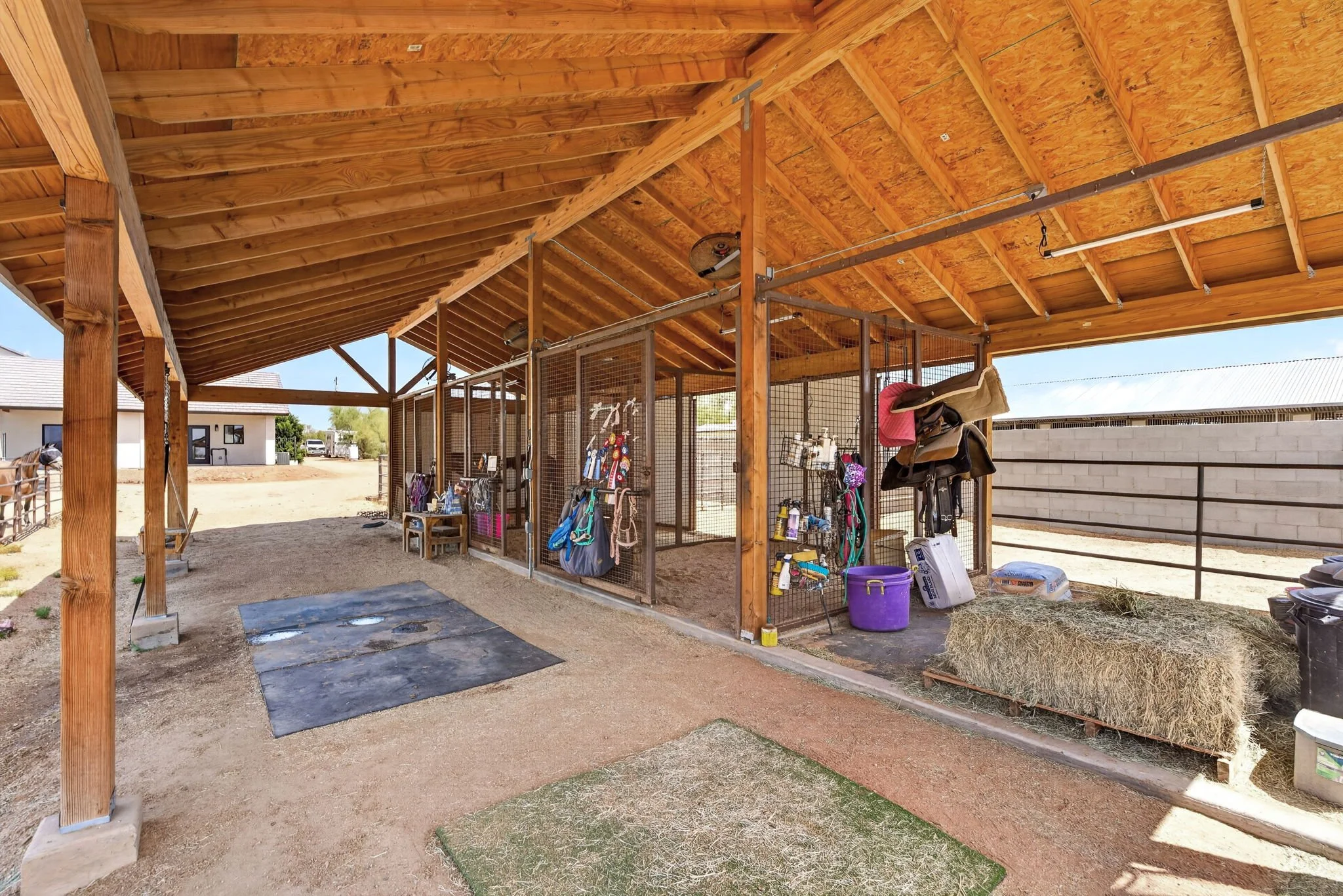 A covered outdoor barn with a wooden roof and support beams, containing horse grooming supplies, tack, and equipment, with a fenced area for horses and a barn or stable building in the background.