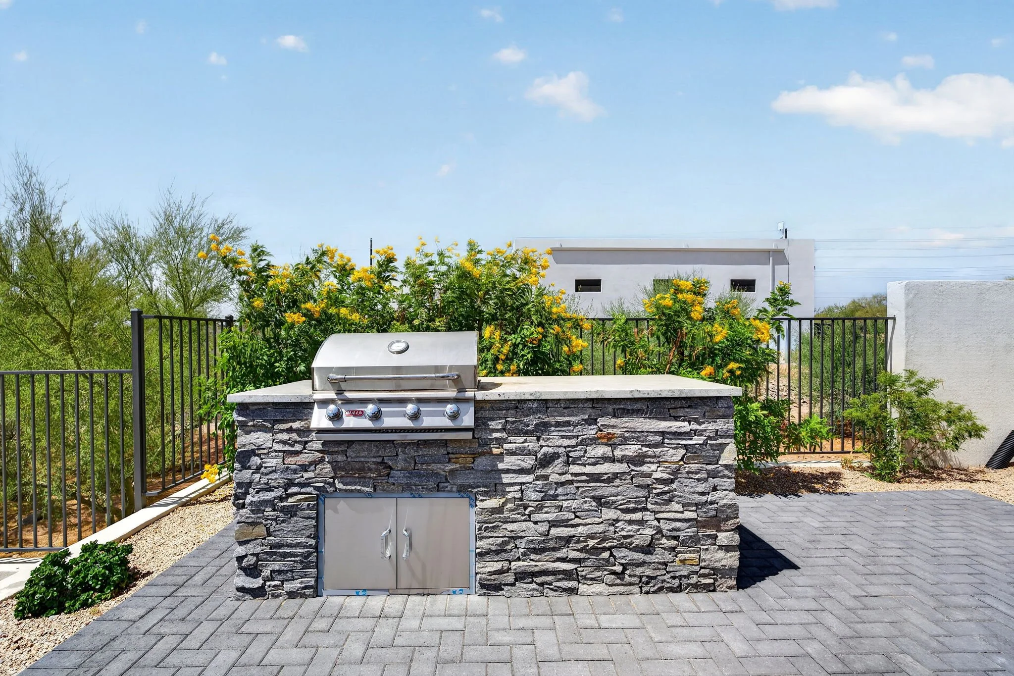 An outdoor patio area with a built-in stainless steel grill on a stone counter, surrounded by a metal fence, with flowering yellow bushes, trees, a building in the background, and a clear blue sky with some clouds.