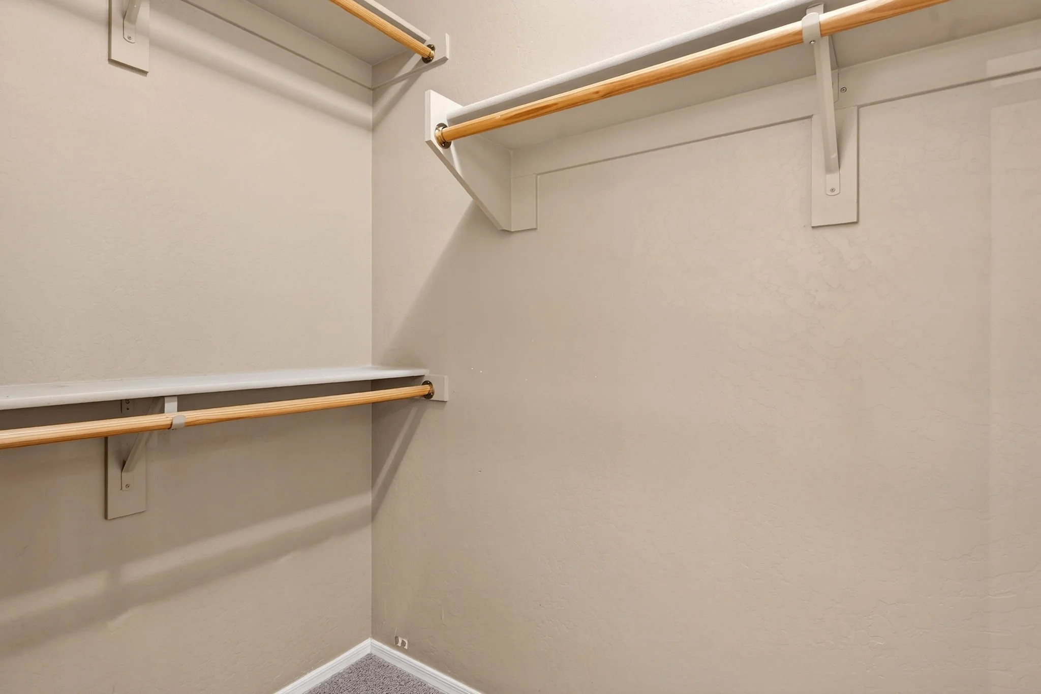 An empty closet with beige walls, gray carpet, and wooden and white shelves with hanging rods.