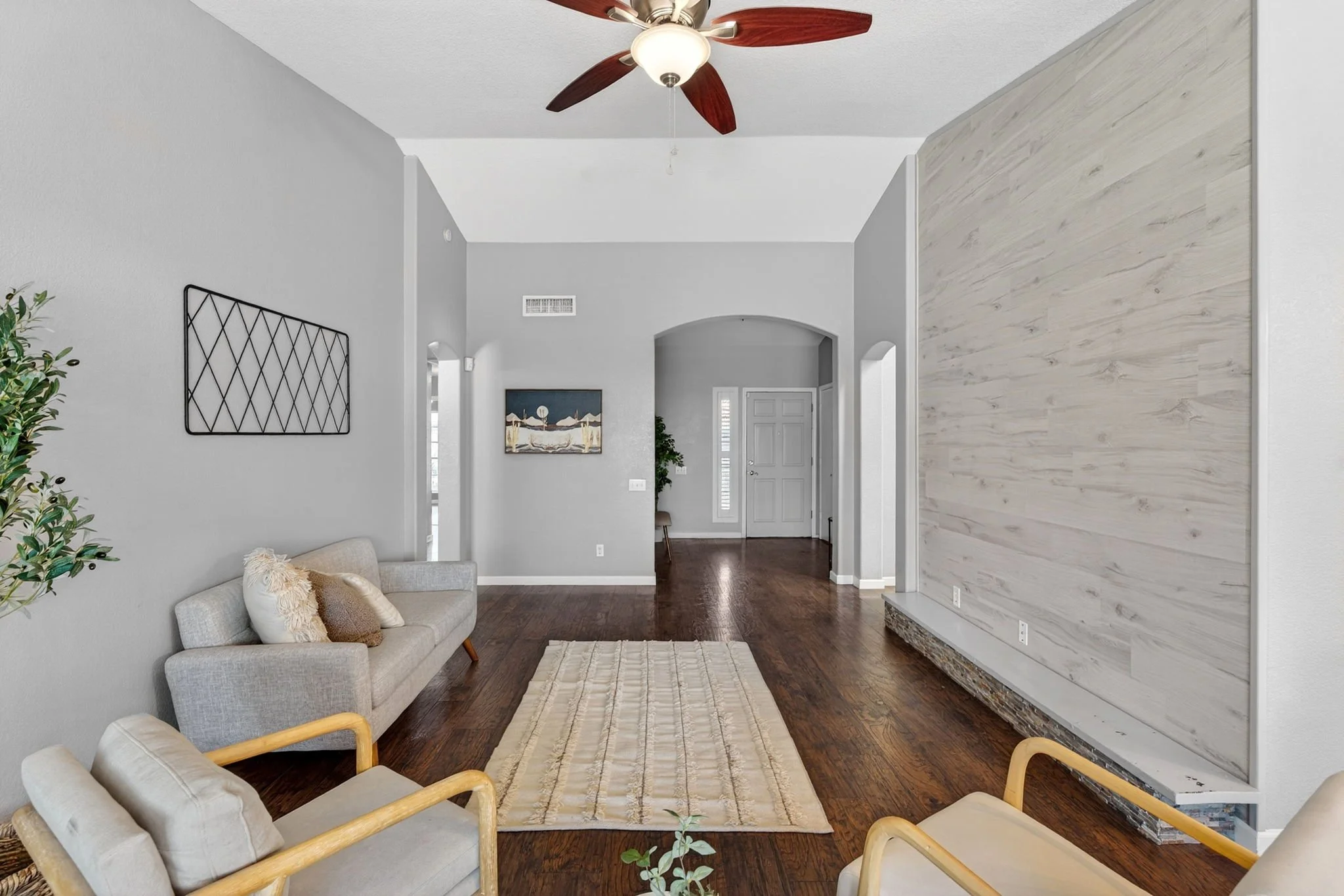 Living room with gray walls, wooden floor, ceiling fan, and a beige sofa with throw pillows. There is a large decorative wood panel on one wall, a framed artwork on the opposite wall, a small plant, and a rug near the sofa.