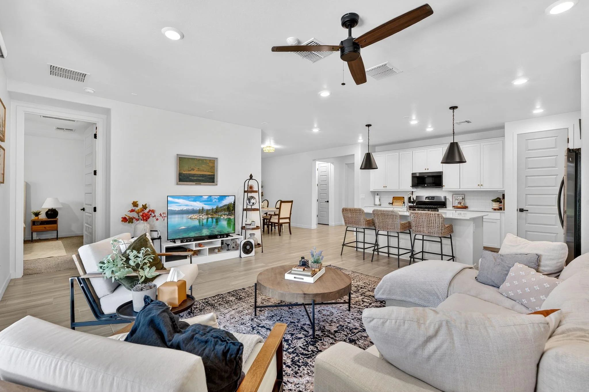 A spacious open-concept living room with white walls, wood flooring, and modern furniture. There is a large white sectional sofa, a black armchair, a round coffee table, and a patterned rug. A flat-screen TV displays a scenic landscape and is placed 