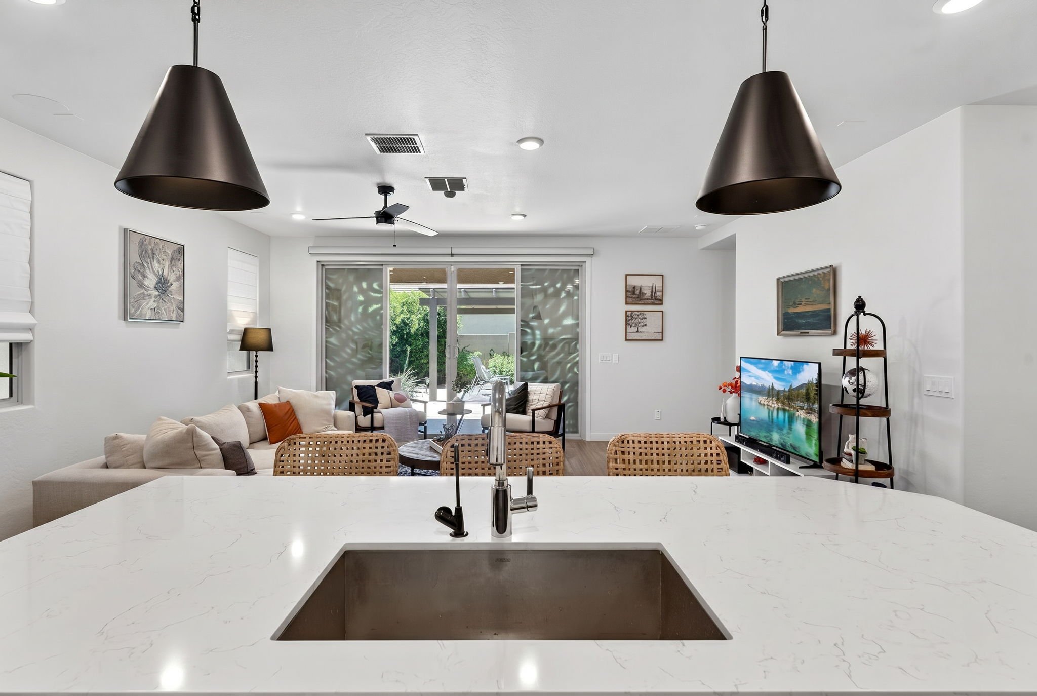 Modern open-concept living room and kitchen area with a white marble kitchen island and sink, beige sofa, wall art, sliding glass door, and a television.